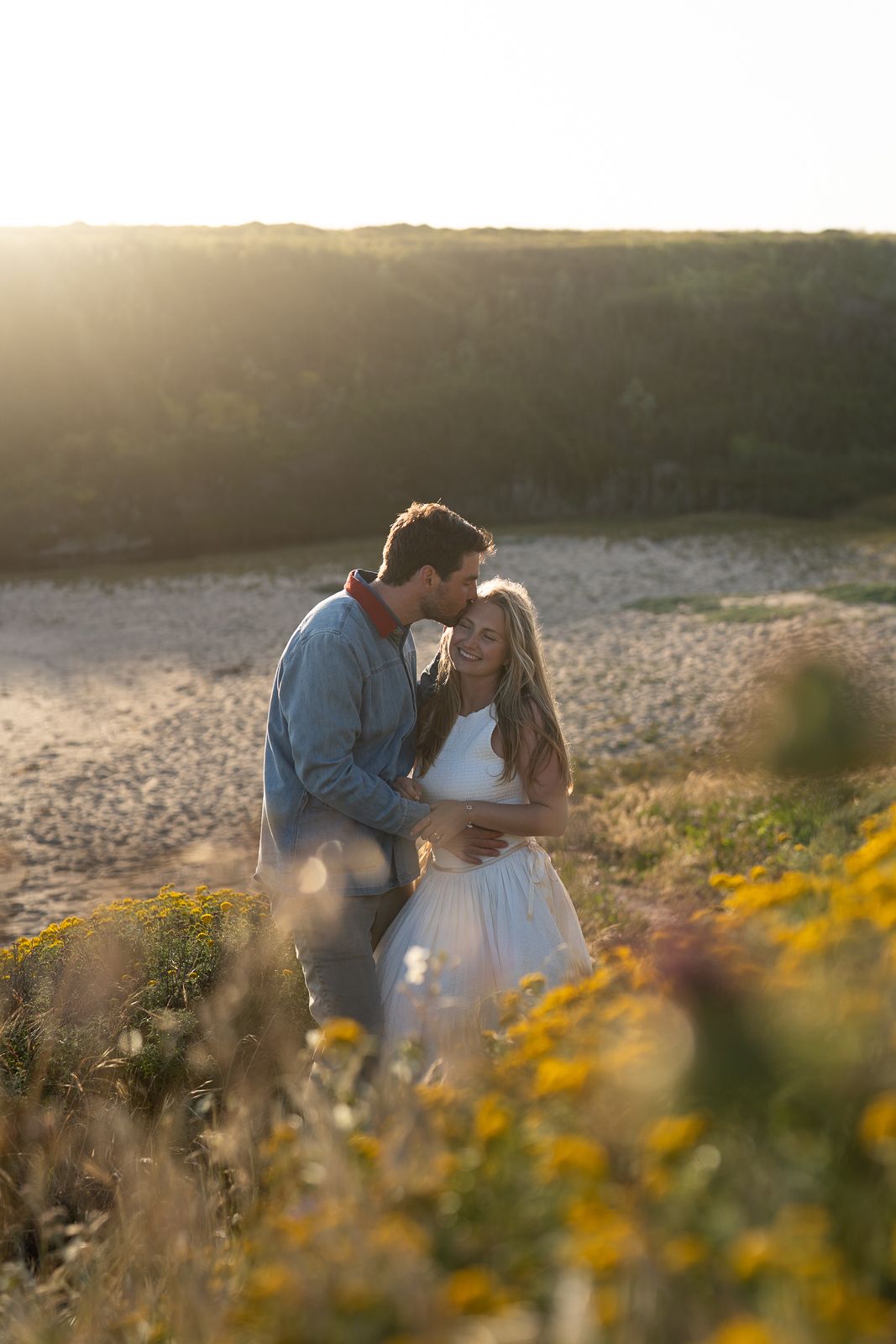 A couple shares a tender moment at sunset among wildflowers in a mountainous landscape.