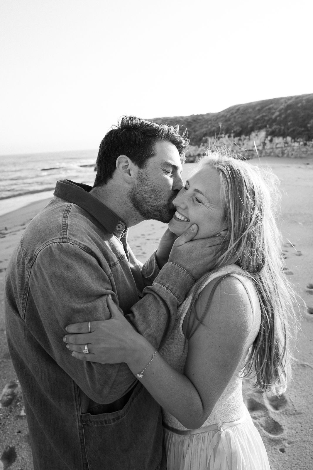 Black and white photo of a romantic couple embracing and laughing together on a beach with cliffs in background.
