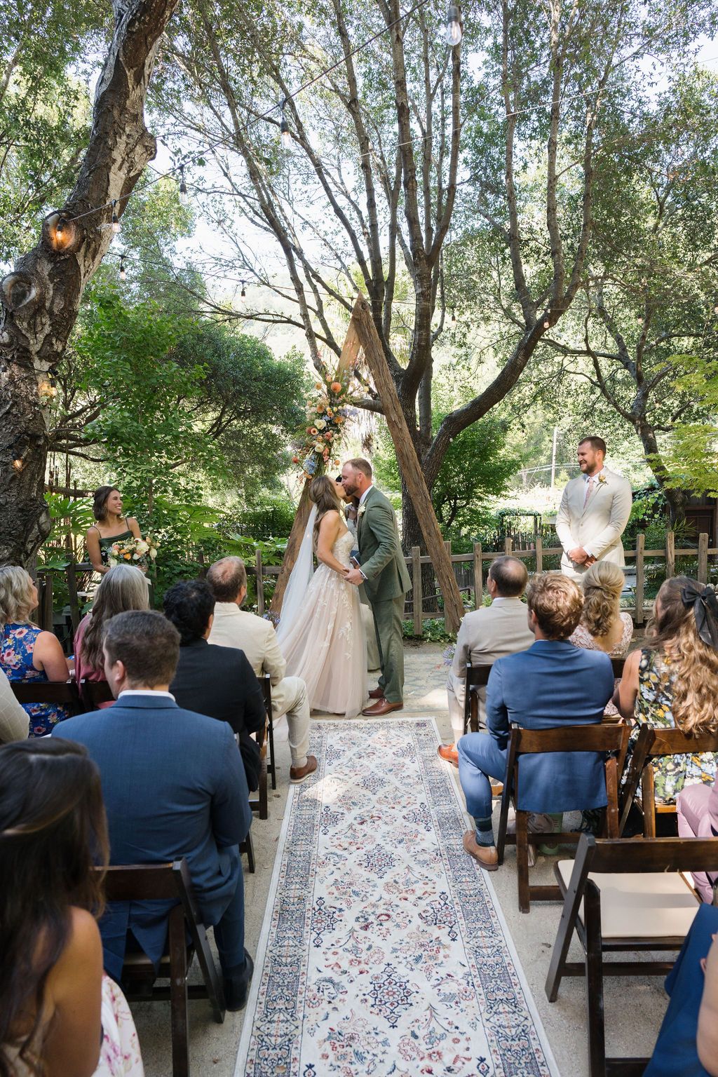A beautiful intimate outdoor garden wedding ceremony with guests seated along a decorative rug aisle under tall trees.