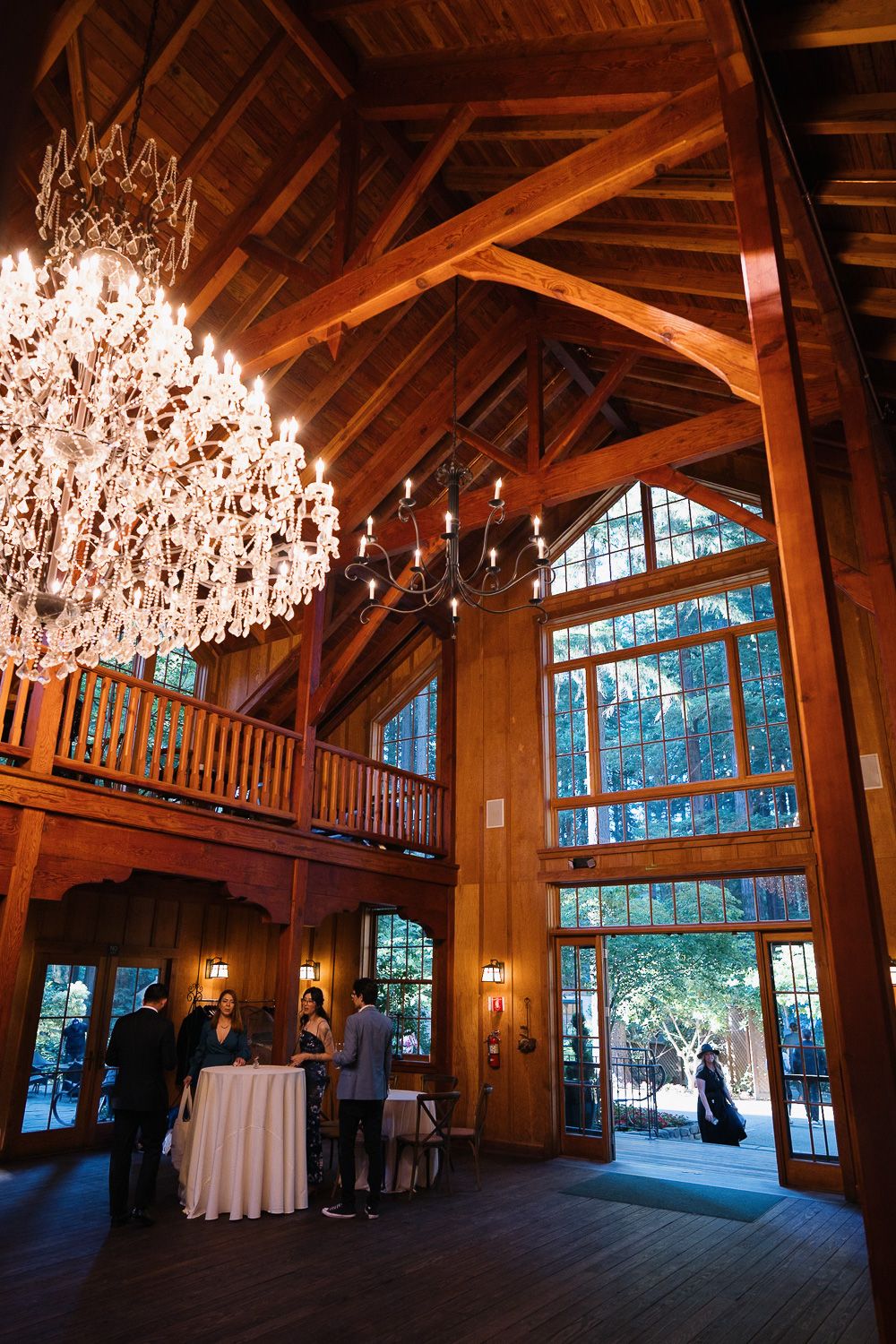 Elegant rustic event venue with vaulted wood beam ceilings, large windows, and sparkling crystal chandeliers in warm lighting.