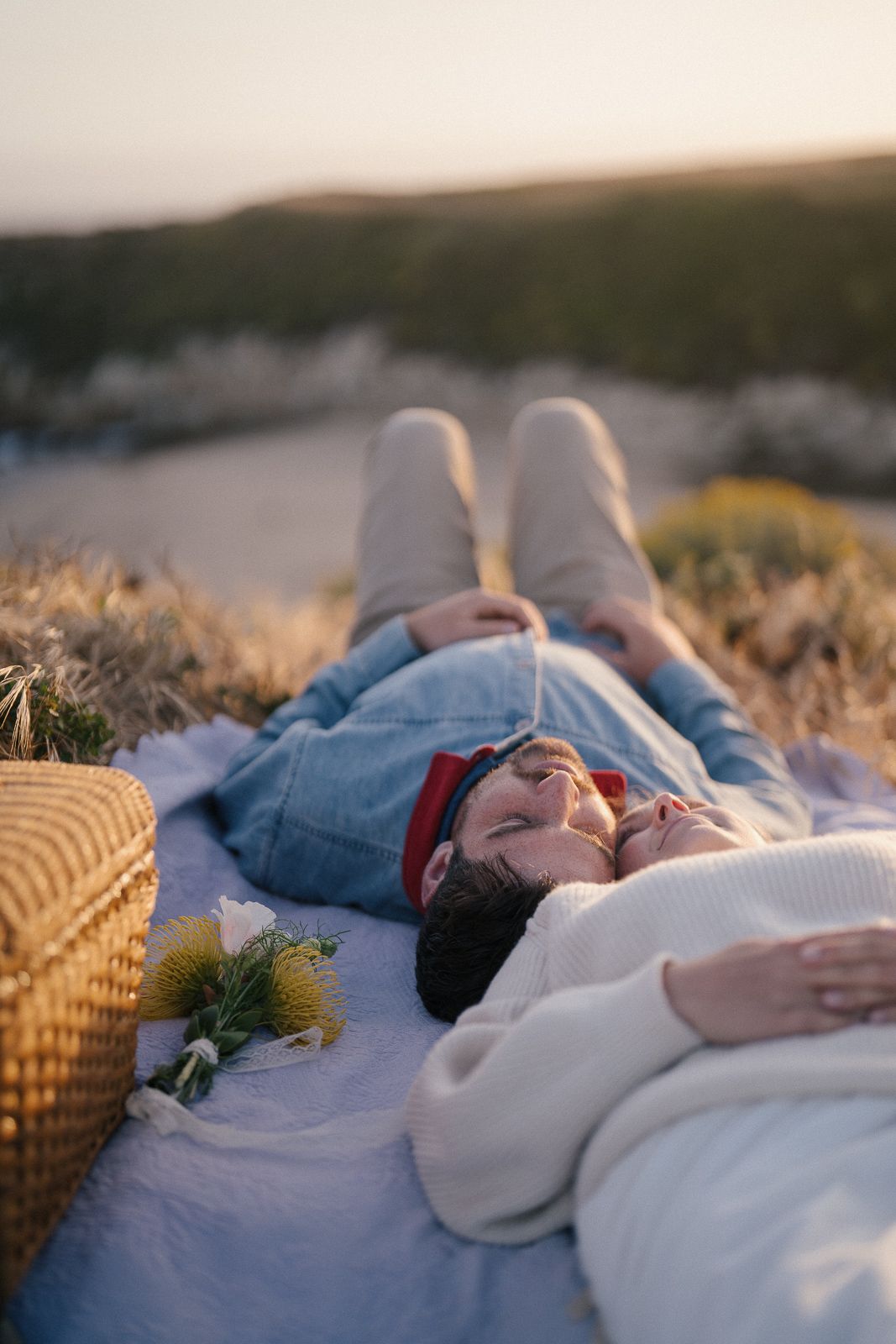 Couple relaxing on picnic blanket at sunset with wicker basket and flowers beside them in golden light.