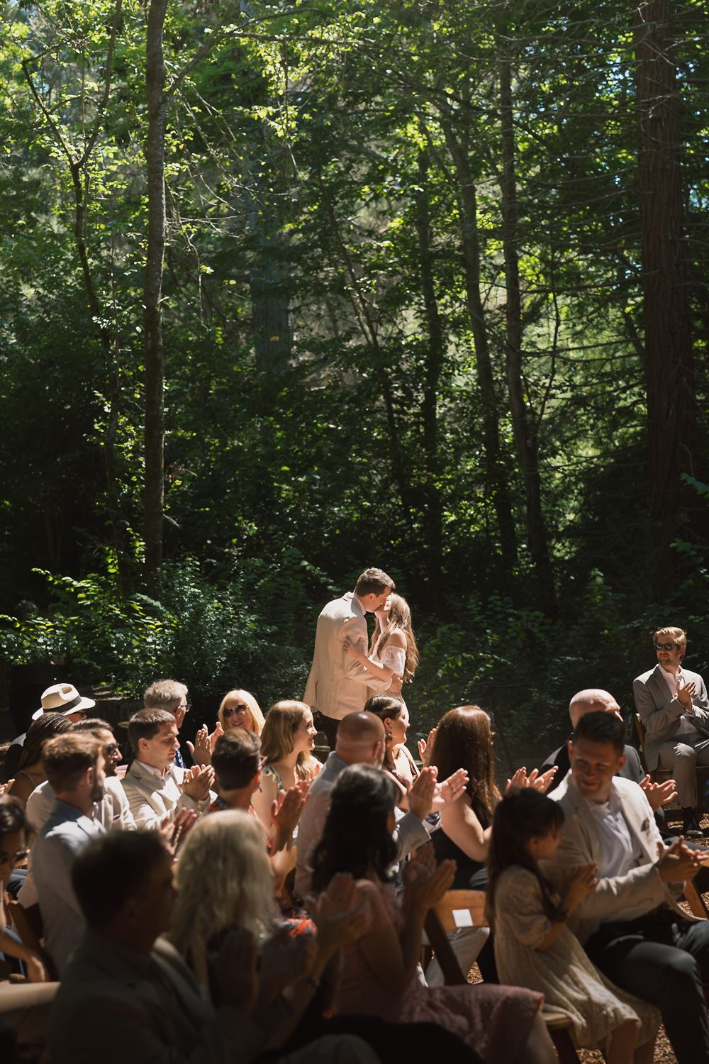 A gathering of people seated outdoors in a sunlit forest clearing during a summer celebration.