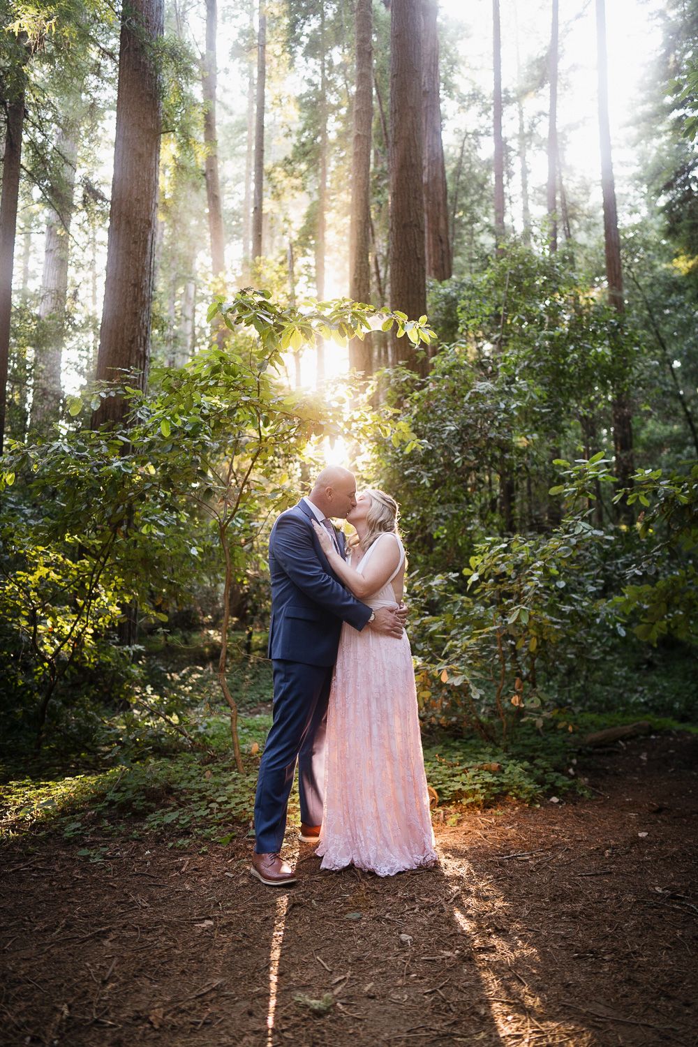 A couple shares a romantic moment in a sunlit forest clearing surrounded by tall evergreen trees and lush greenery.