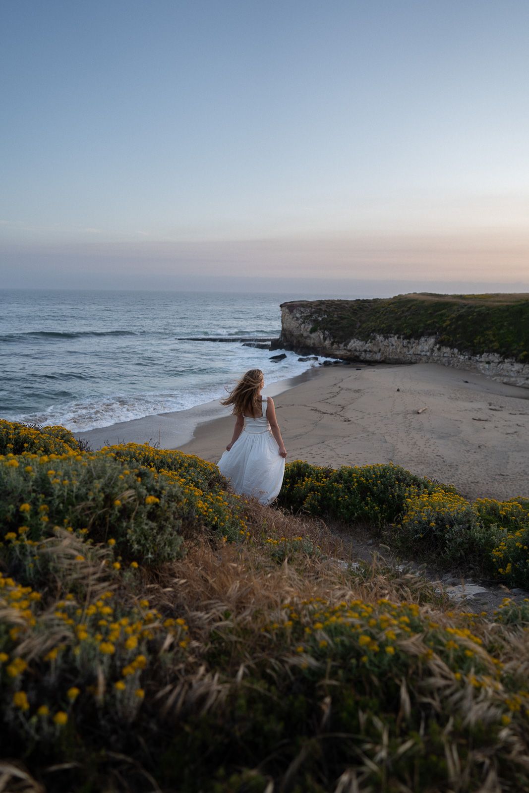 Person in white dress stands on coastal cliff overlooking sandy beach with yellow wildflowers during sunset.