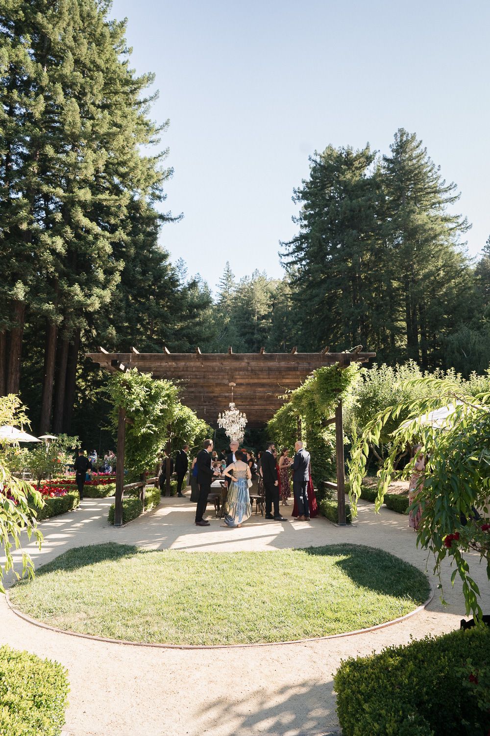 Outdoor wedding ceremony setup with wooden arbor and curving path surrounded by evergreen trees and lush landscaping.
