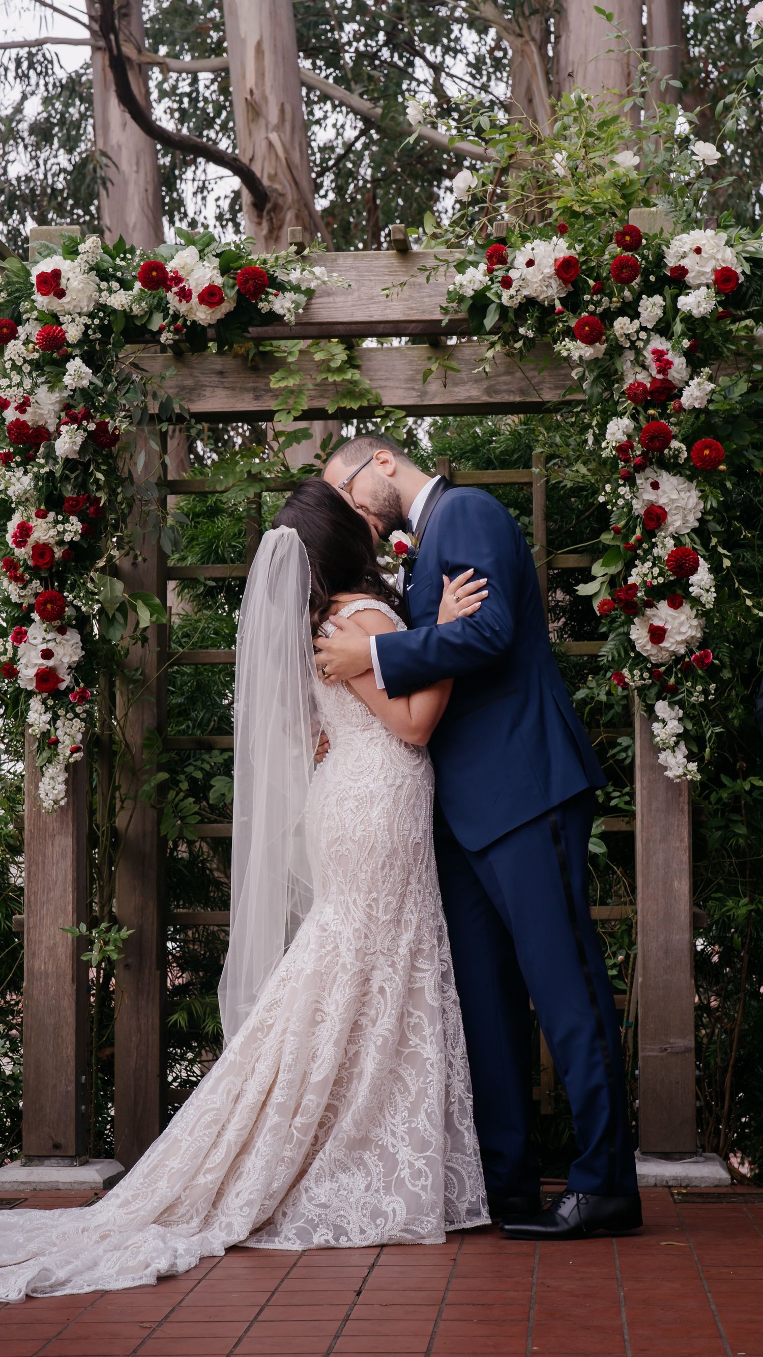 Romantic wedding ceremony under wooden arch decorated with red and white roses on brick patio surrounded by eucalyptus trees.