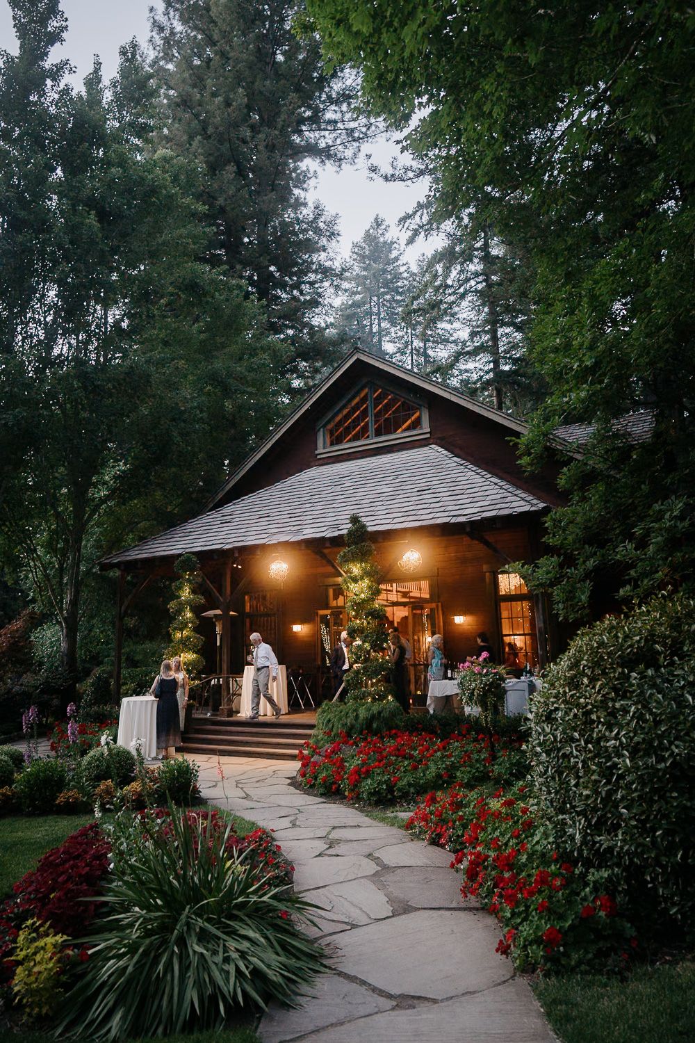Rustic wooden cabin venue illuminated by warm porch lights with stone pathway and vibrant red flowers at dusk.