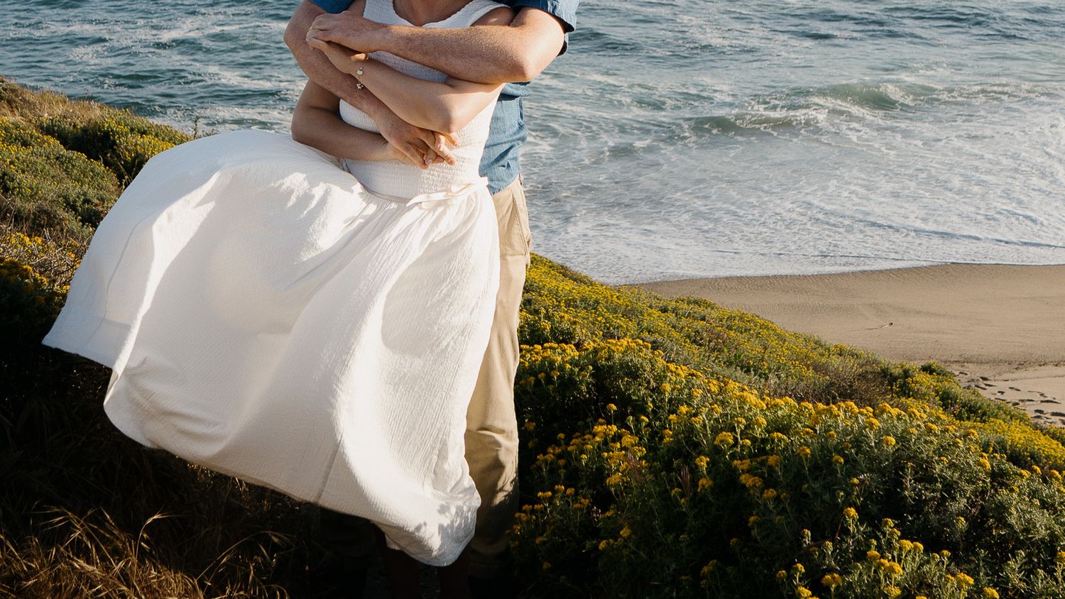 A white wedding dress flows in the breeze on a coastal cliff overlooking the ocean with yellow wildflowers in bloom.