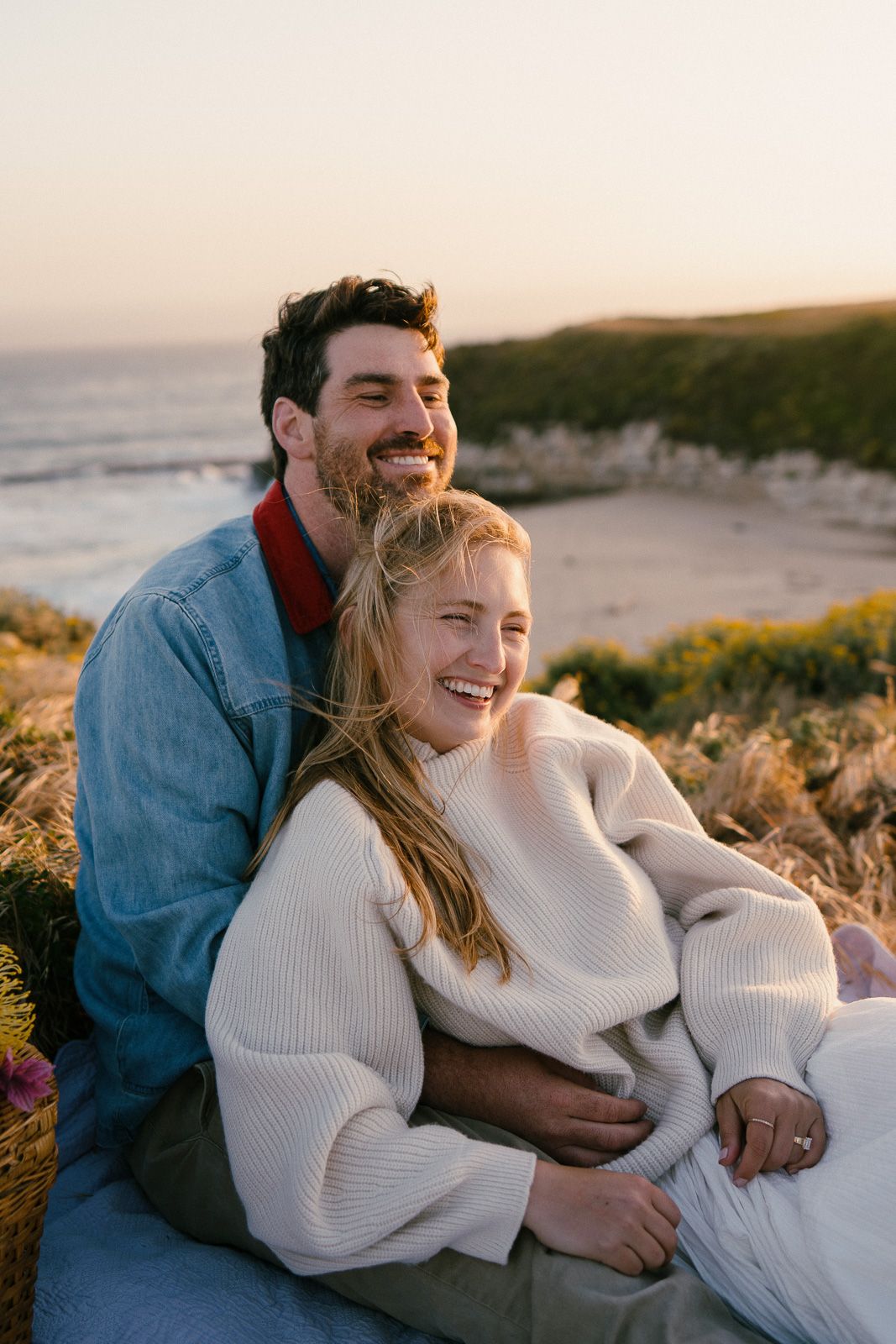 A couple shares a joyful moment while sitting on coastal cliffs at sunset, wearing cozy sweaters by the ocean.