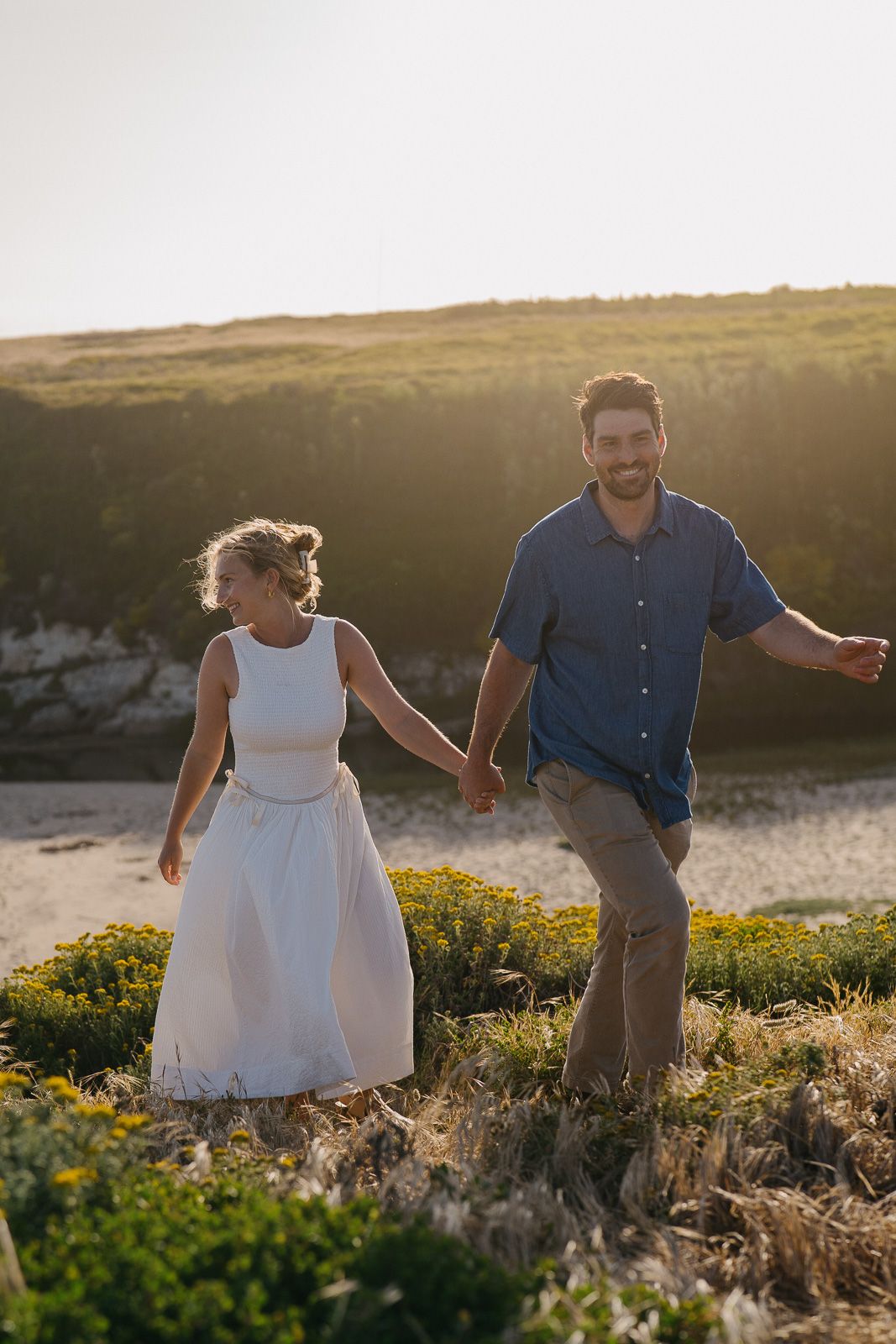 Couple walking through wildflowers on a hilltop at golden hour holding hands in romantic countryside setting.