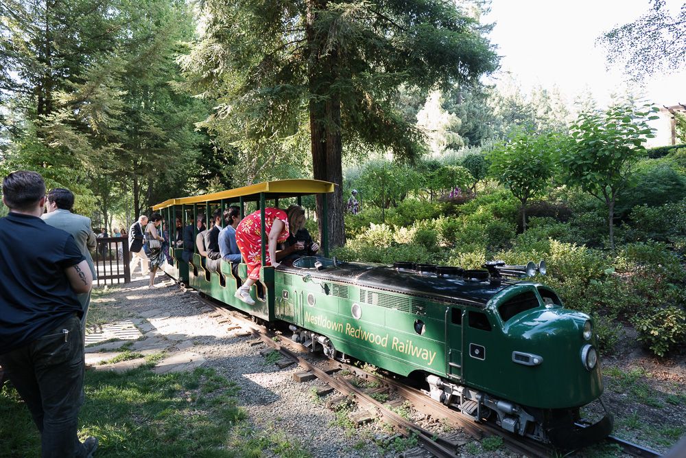 A miniature green train carries passengers through a lush garden setting on a sunny day.