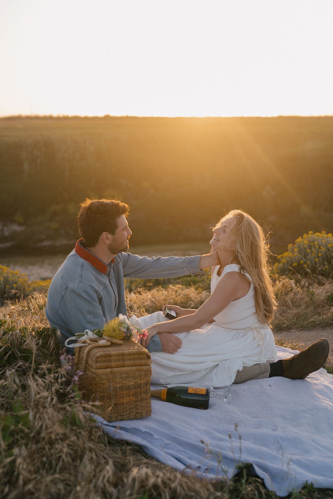 Couple having romantic sunset picnic on blanket with wine basket in golden field during summer evening.