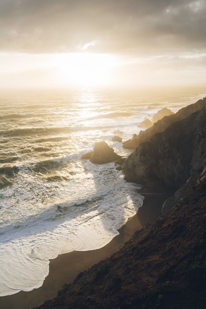 Dramatic coastal sunset view with golden light illuminating rocky cliffs and waves crashing on a secluded beach.