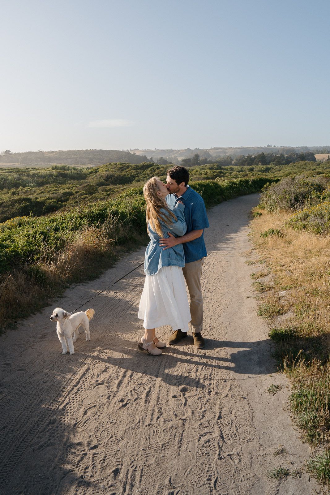 Couple shares a tender moment on a coastal hiking trail with their white dog on a sunny summer afternoon.