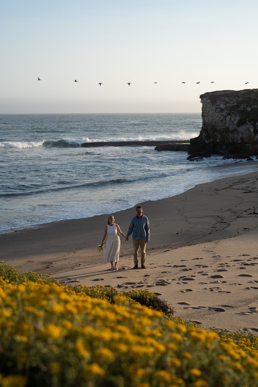 Two people walk along a beach at sunset with cliffs, ocean waves and yellow wildflowers in California.