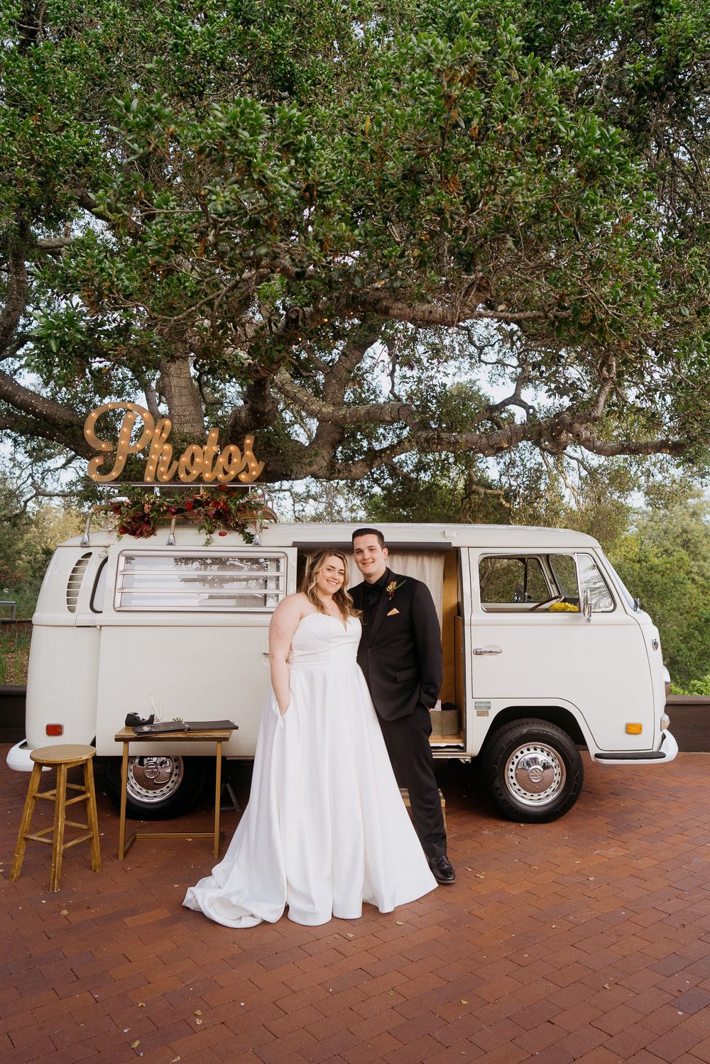 A vintage white VW van with a retro photo booth setup parked outdoors under lush green trees.