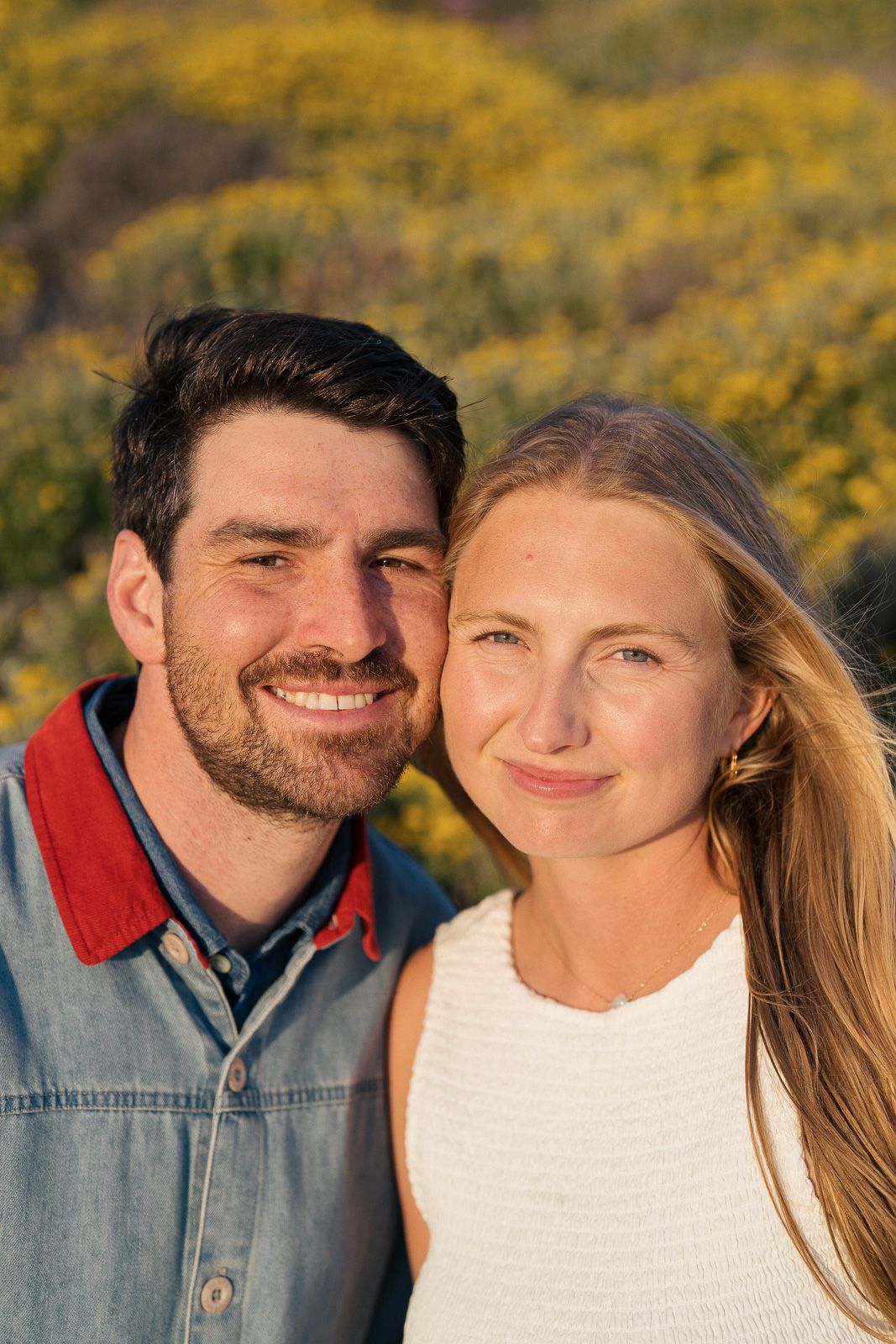 A happy couple smiles outdoors on a sunny day with yellow flowers in the background.