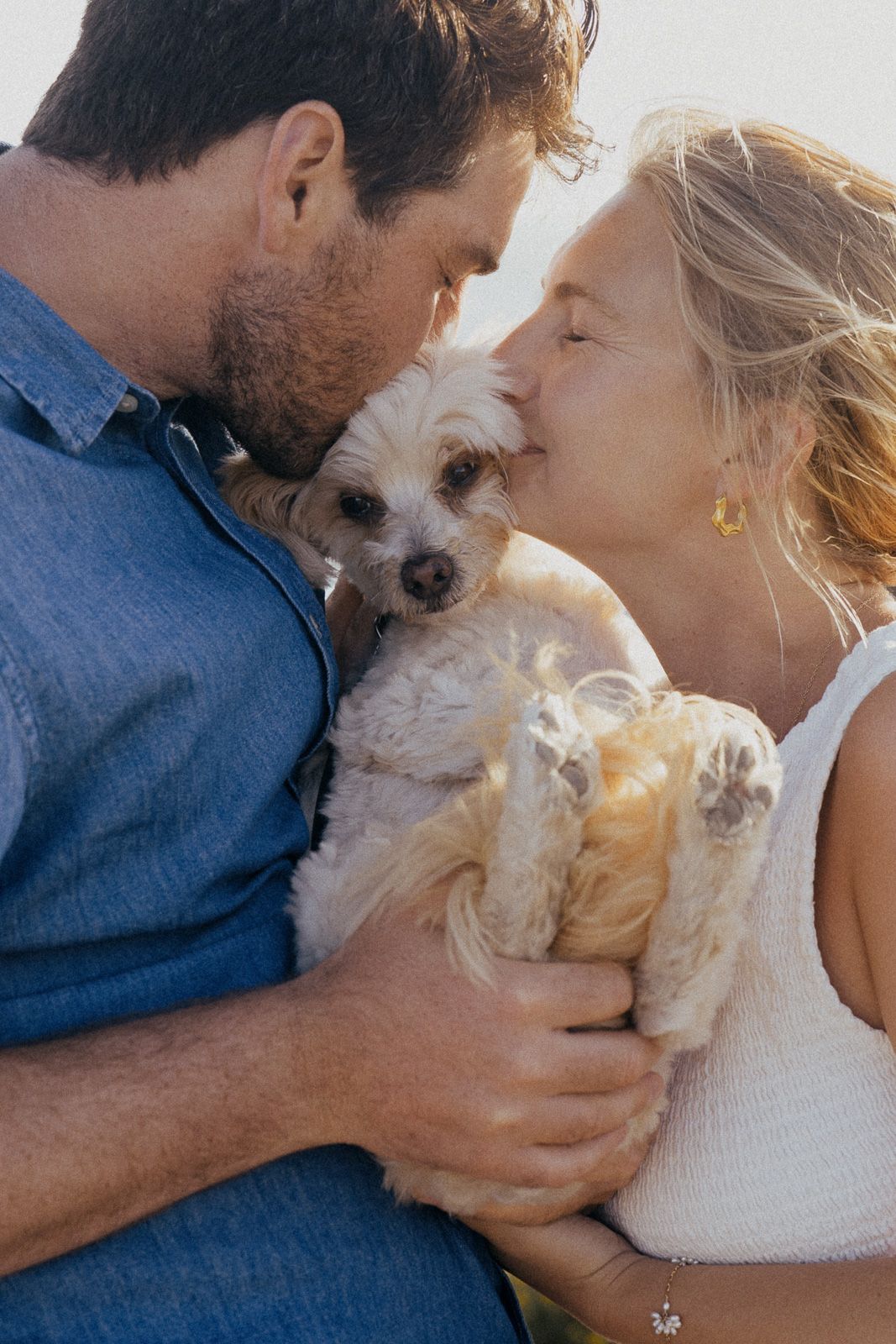 A couple shares a tender moment with their small white fluffy dog in a sunlit outdoor setting.