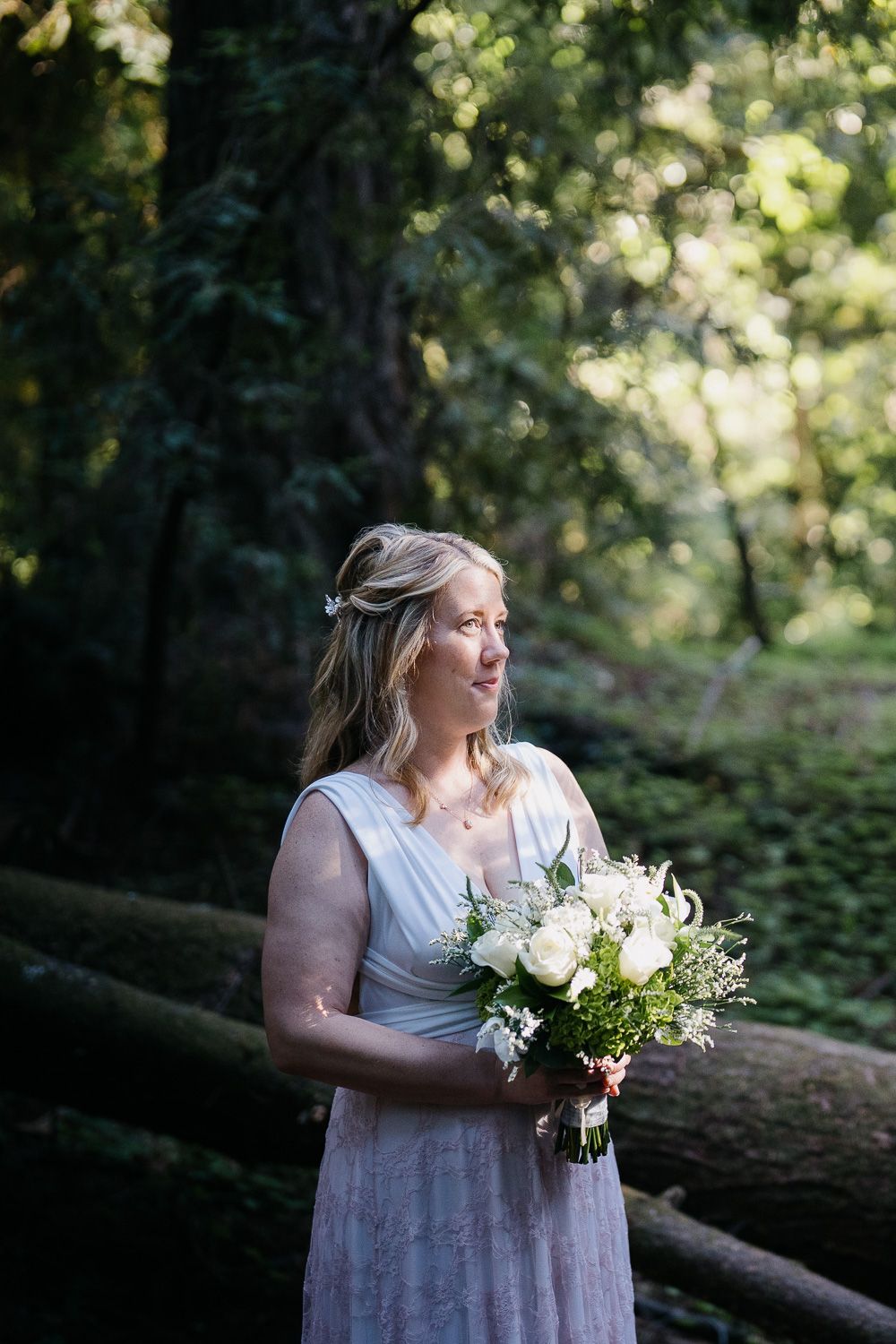 A bride in a light flowy dress holds a white floral bouquet while standing in a scenic wooded forest setting.
