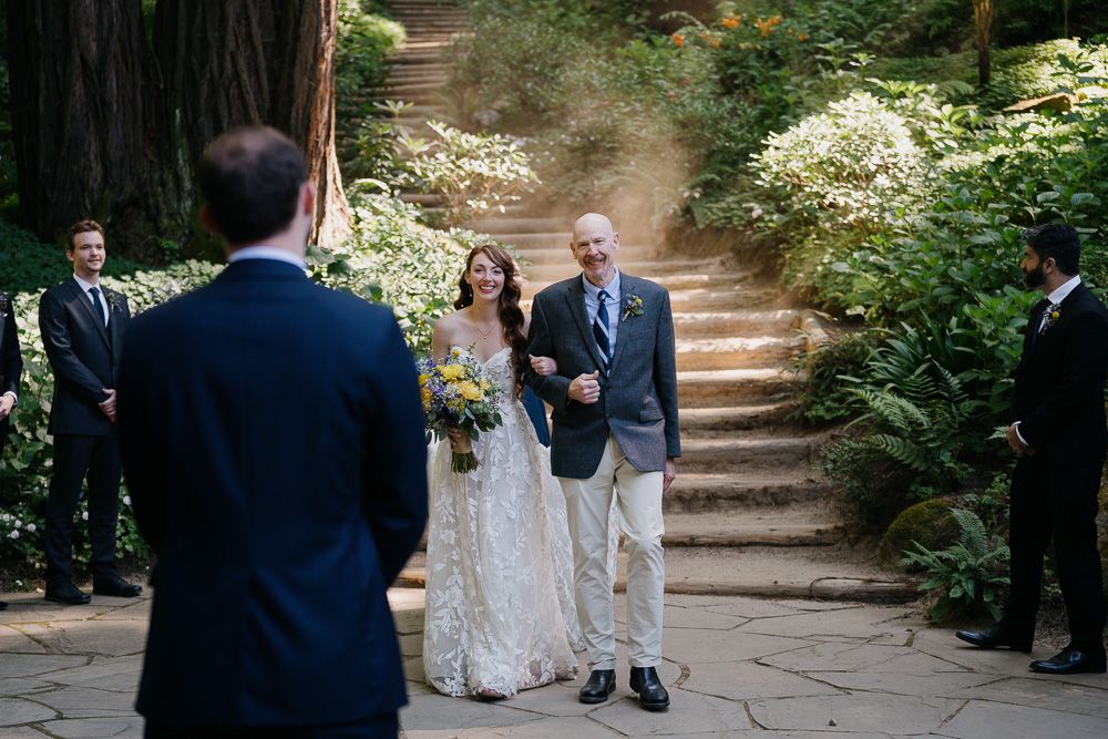 A beautiful outdoor wedding ceremony takes place on rustic stone steps surrounded by lush greenery and flowering plants.