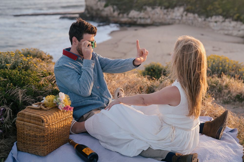 A romantic picnic scene at sunset on a coastal cliff with a wicker basket and blanket spread out by the ocean.