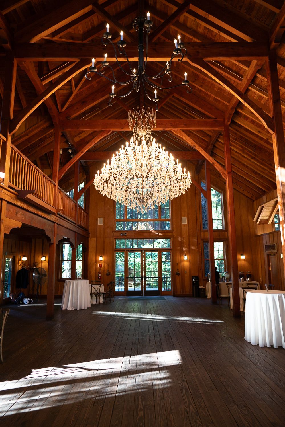 Elegant wooden barn venue interior with exposed beams, crystal chandelier and sunlight streaming through glass doors.