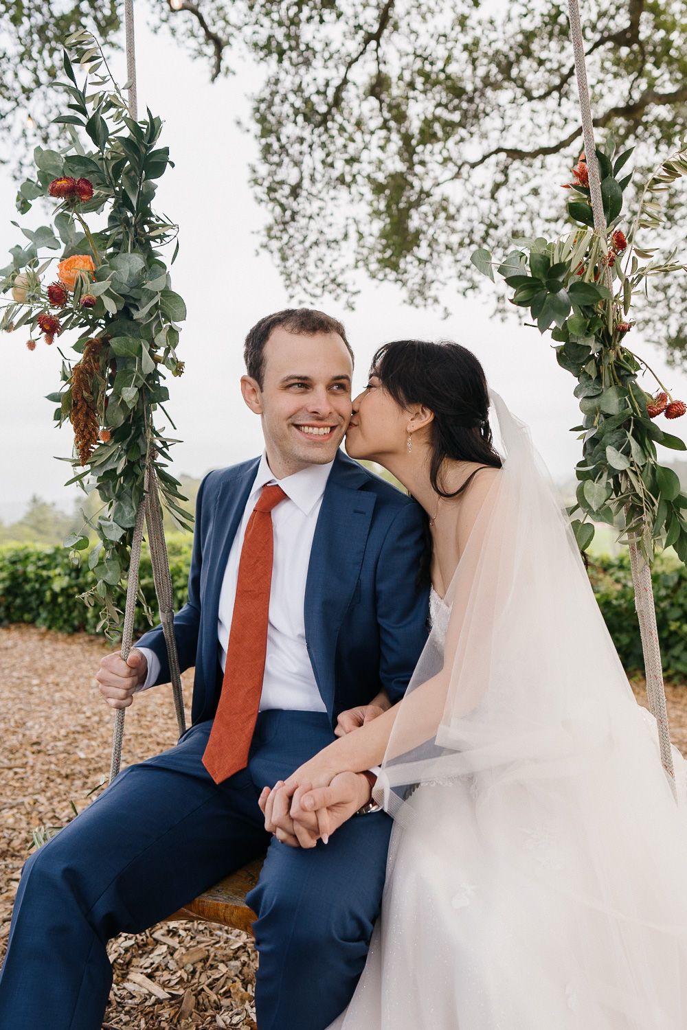 A couple sits on a swing decorated with greenery and flowers at their outdoor wedding celebration.