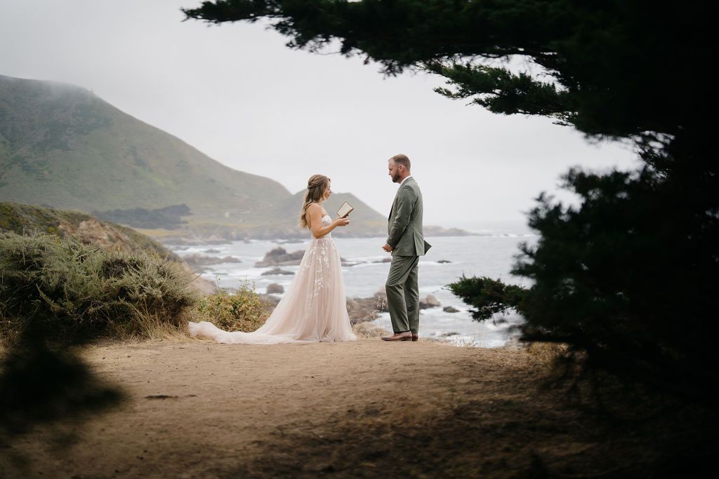 A couple stands at a scenic coastal overlook with dramatic cliffs and ocean waves along a misty California shoreline.