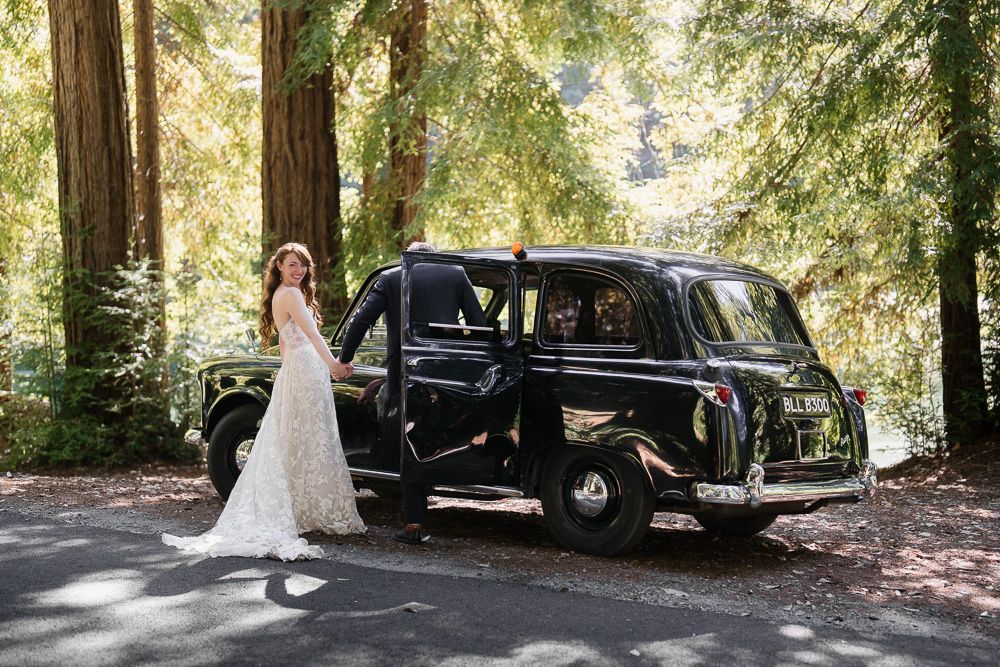 A classic black vintage car parked on a forested road with someone in a white wedding dress standing beside it.