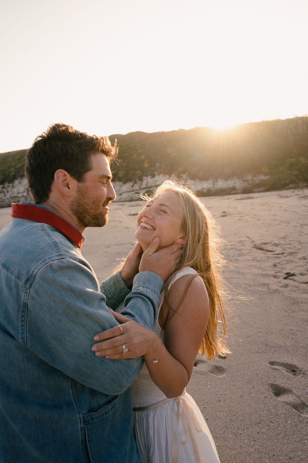 A romantic couple shares an intimate moment on the beach at sunset, dressed in casual attire.