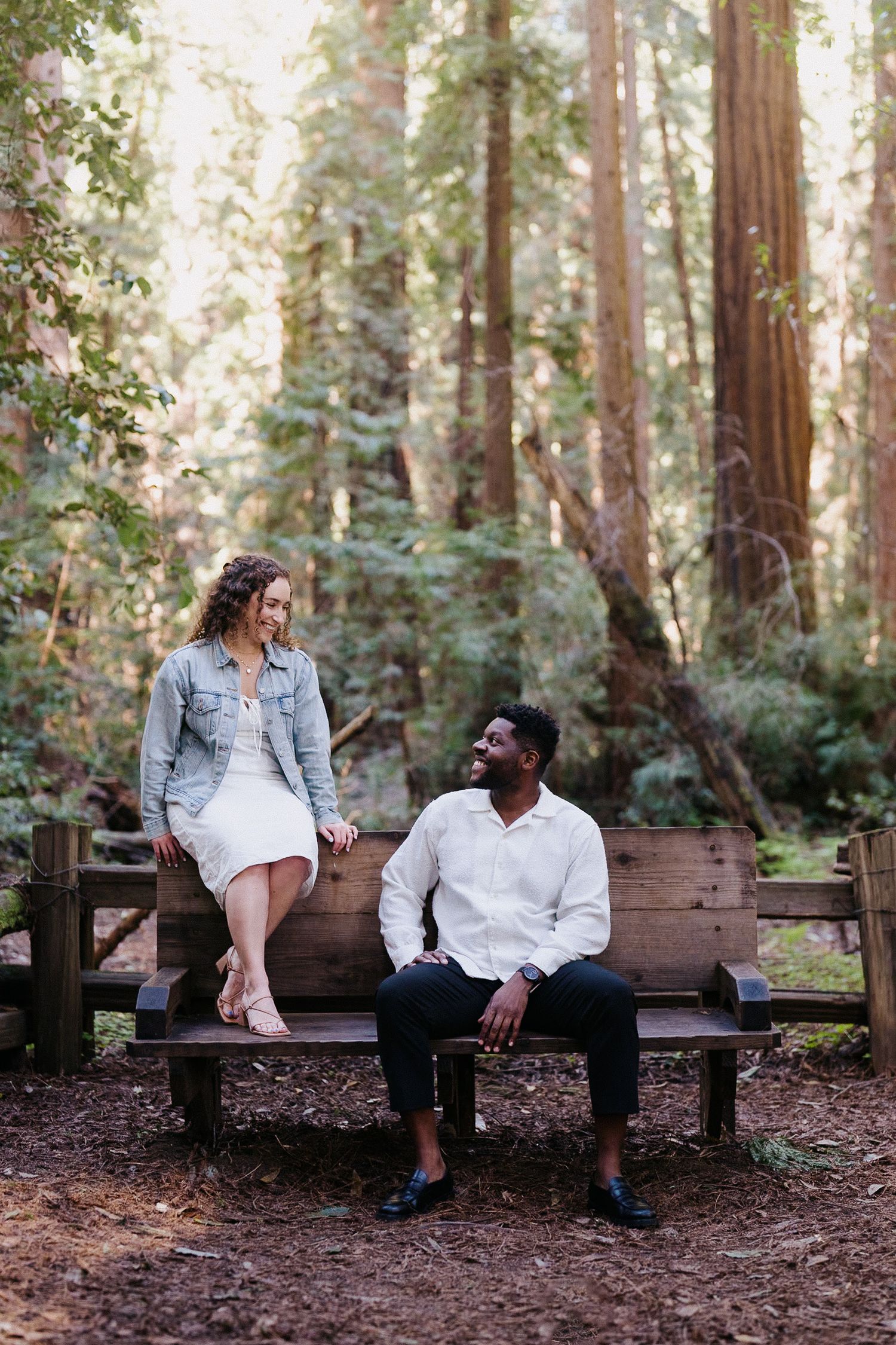 Two people sitting at opposite ends of a wooden bench in a serene redwood forest setting.