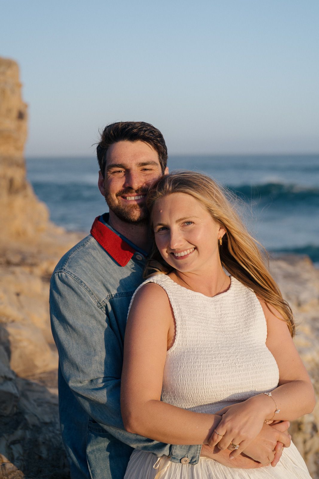 A couple embraces by the ocean at golden hour, with rocky cliffs in the background.