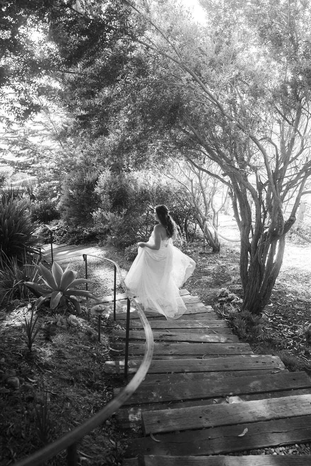 A figure in a flowing white dress runs gracefully across a wooden boardwalk path through a lush garden setting.