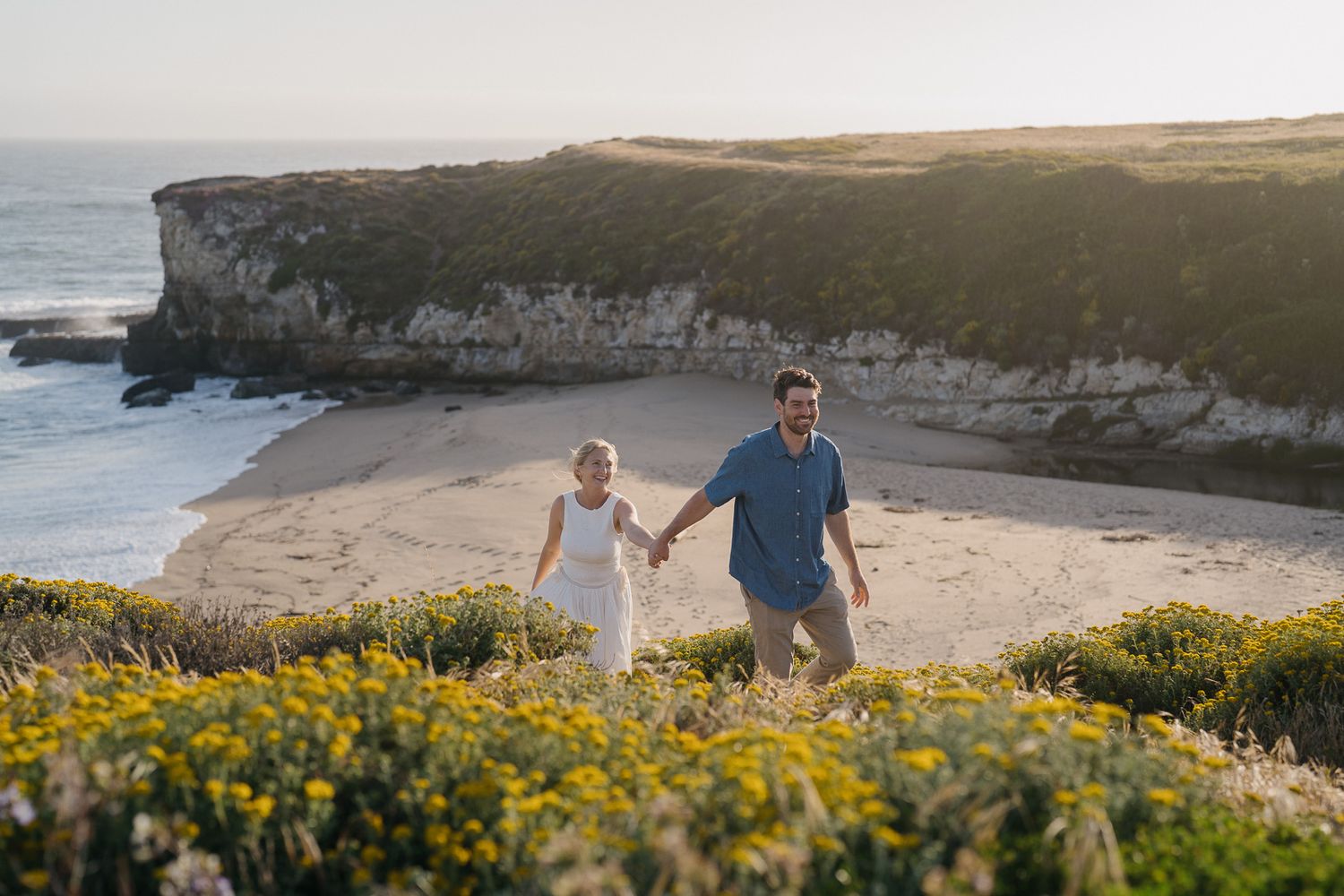Couple exploring coastal cliffs with beach views while walking through yellow wildflowers at sunset.