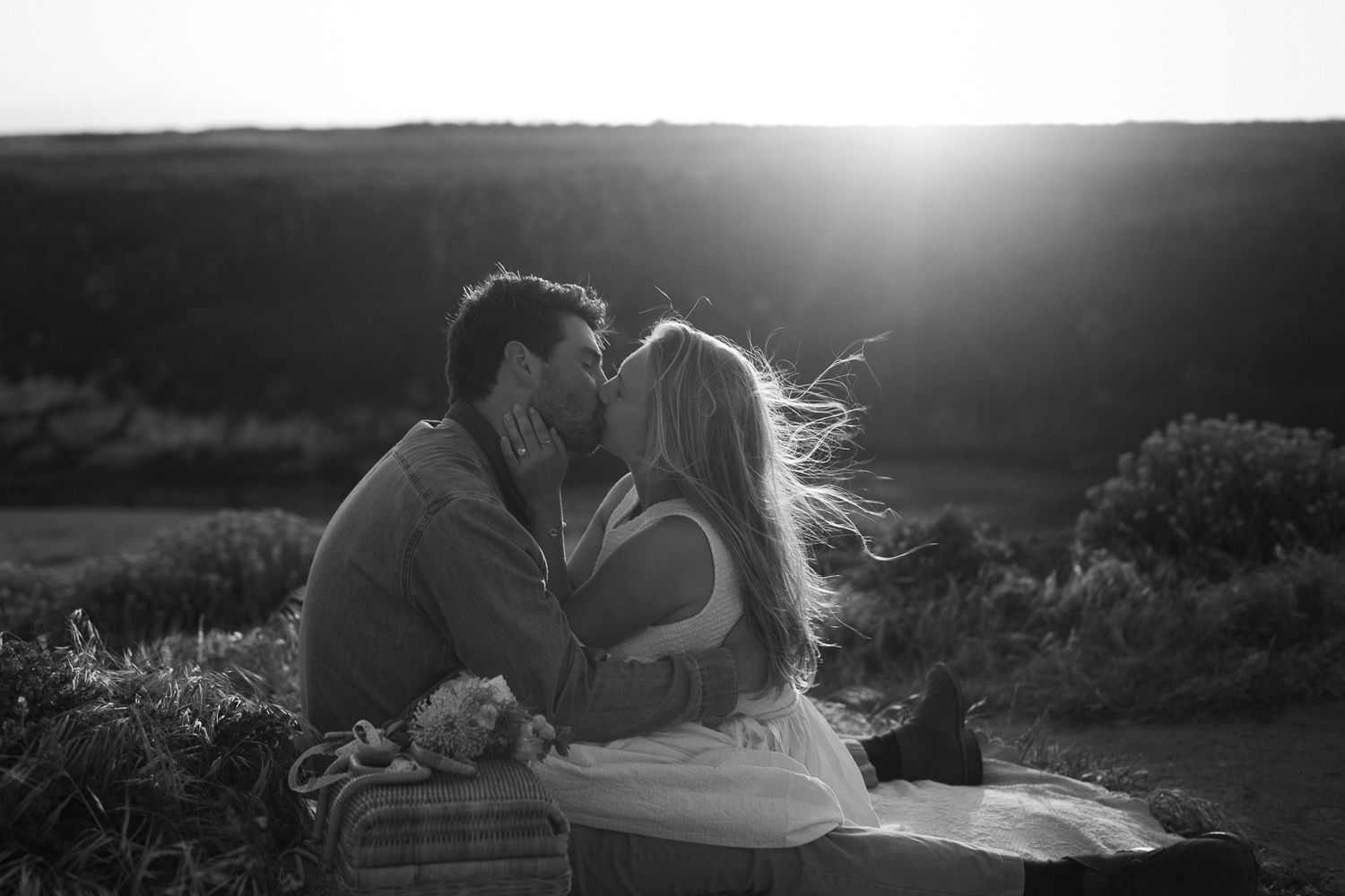A couple shares a romantic black and white moment at sunset on a hilltop with sun flare in the background.