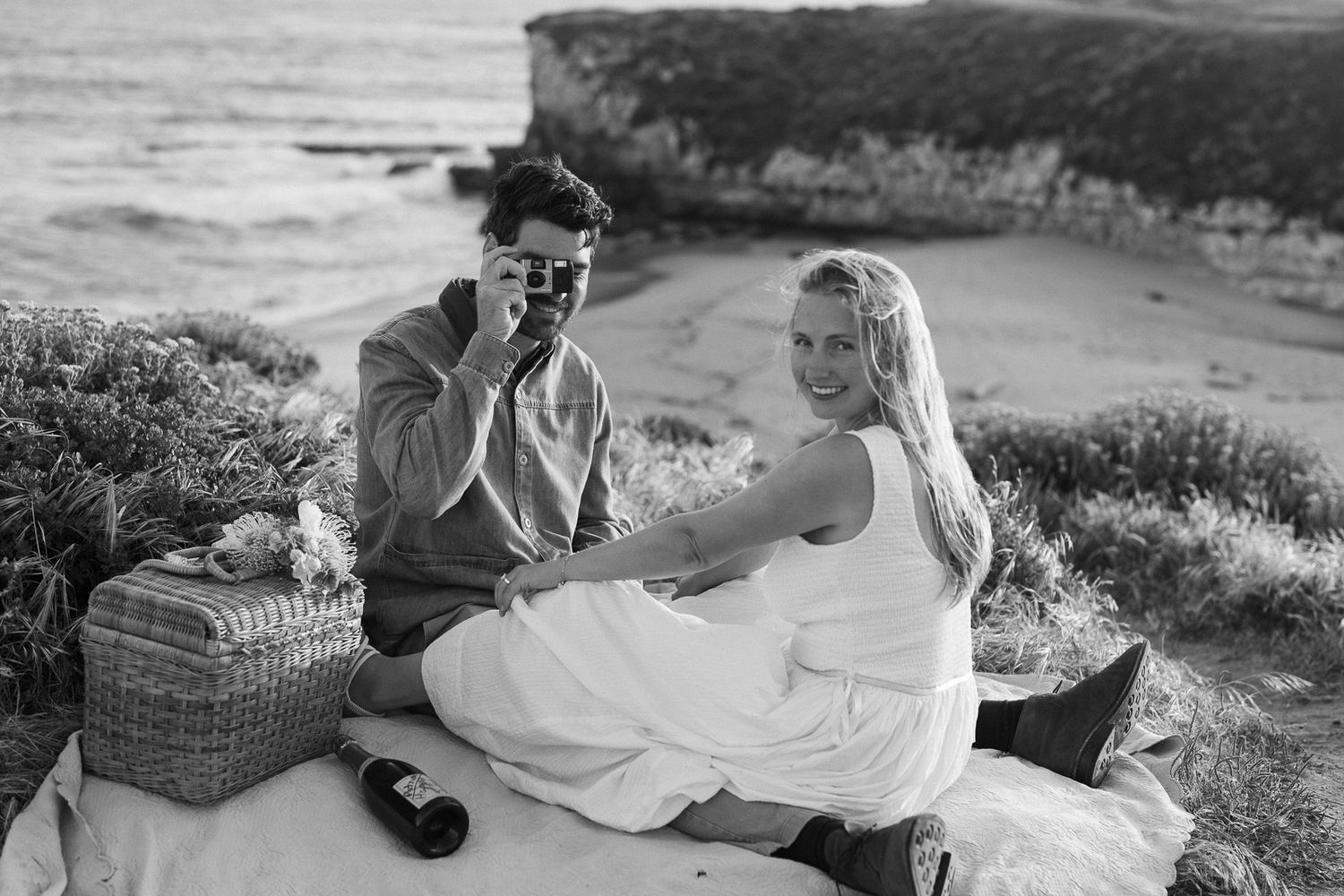 A romantic black and white picnic moment on a coastal cliff overlooking a scenic beach and ocean vista.