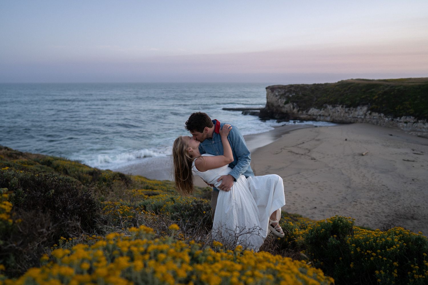 A romantic couple embraces on a coastal cliff overlooking the ocean at sunset, surrounded by yellow wildflowers.