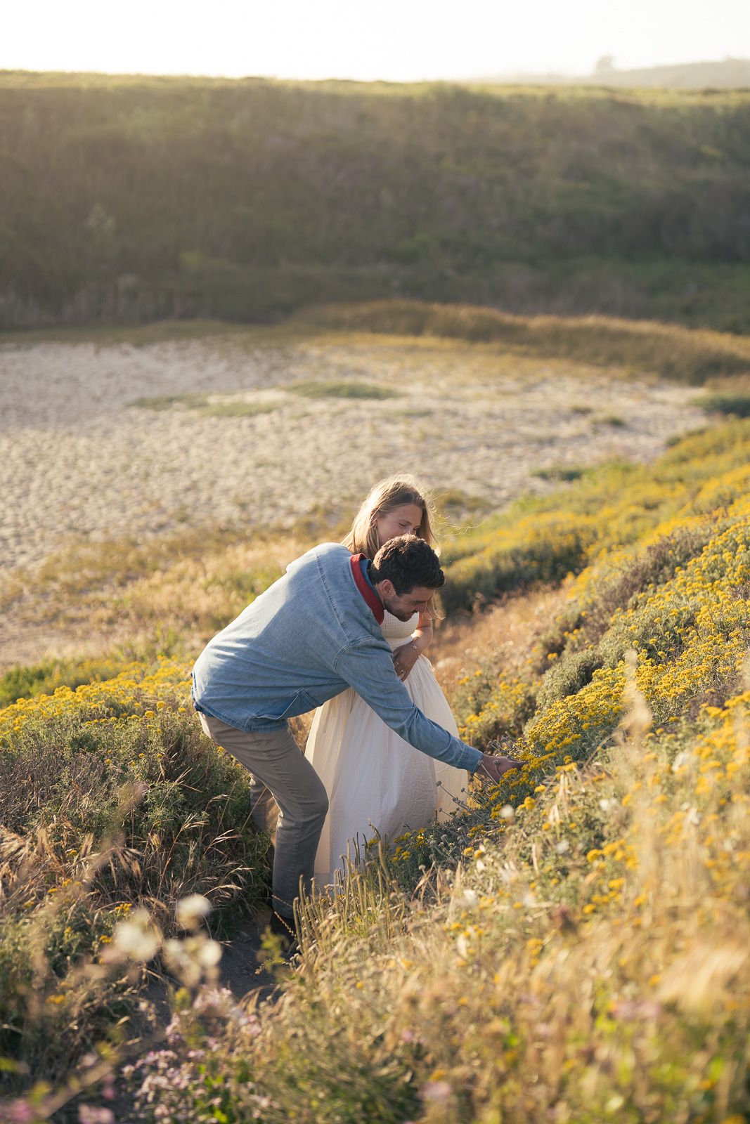 Person in a light gray sweater picking yellow wildflowers in a sunny meadow during golden hour.