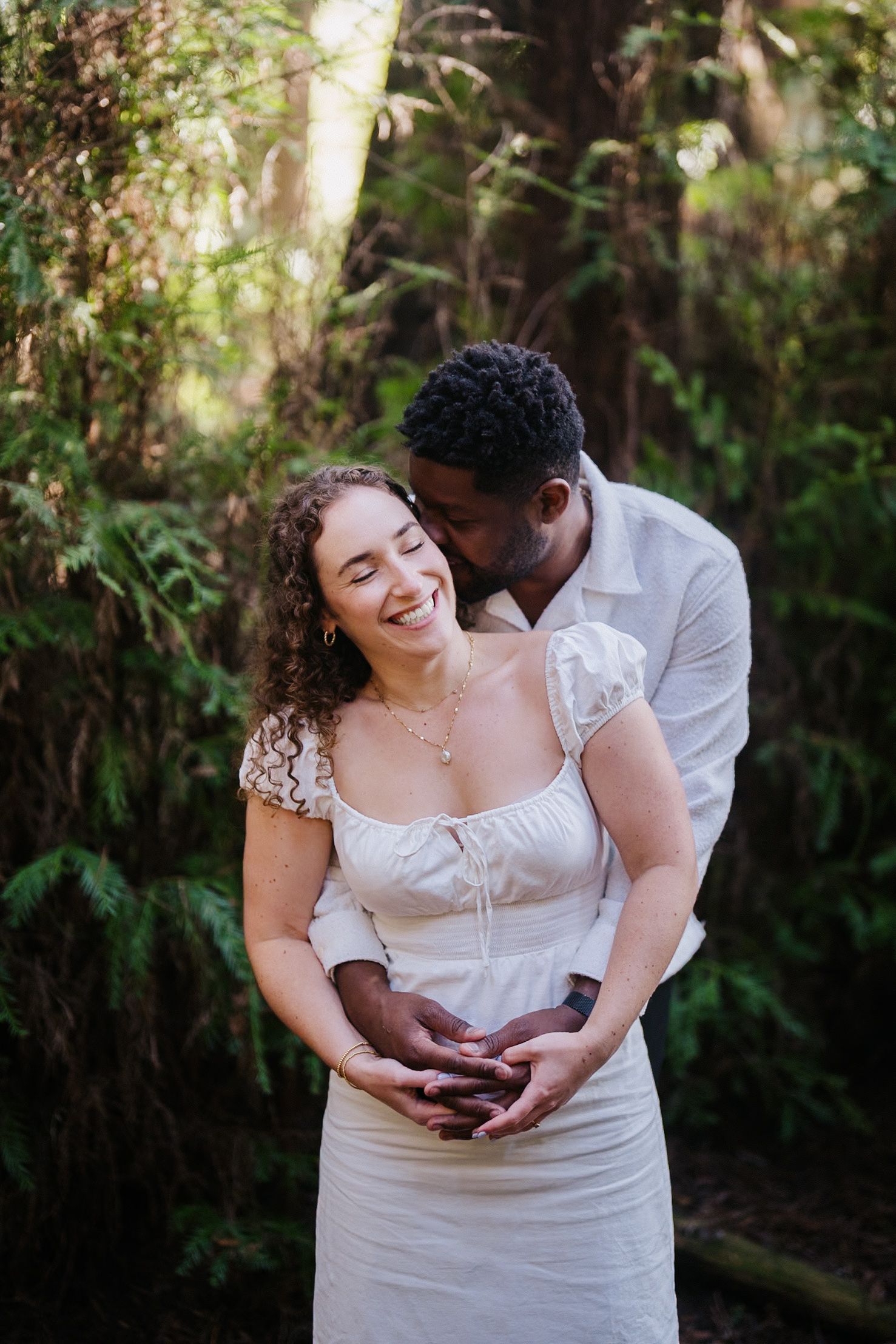 Joyful couple embraces in a lush garden setting as sunlight filters through the trees.