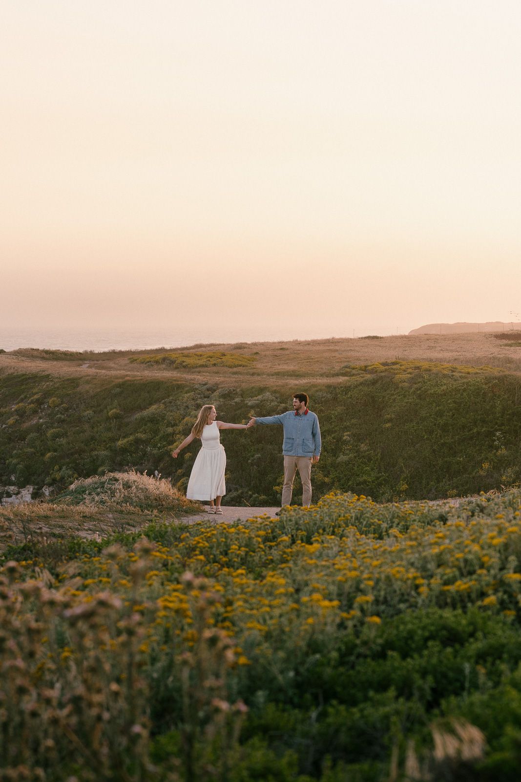 A couple stands together in a field of yellow wildflowers during golden hour on rolling coastal hills at sunset.