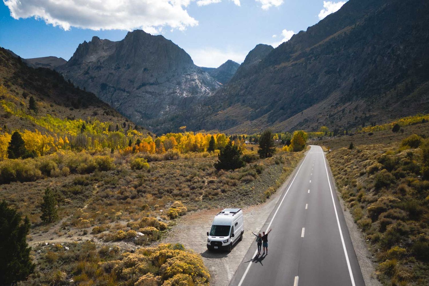 A white vehicle drives on a mountain road surrounded by autumn foliage and rugged peaks in a scenic mountain valley.