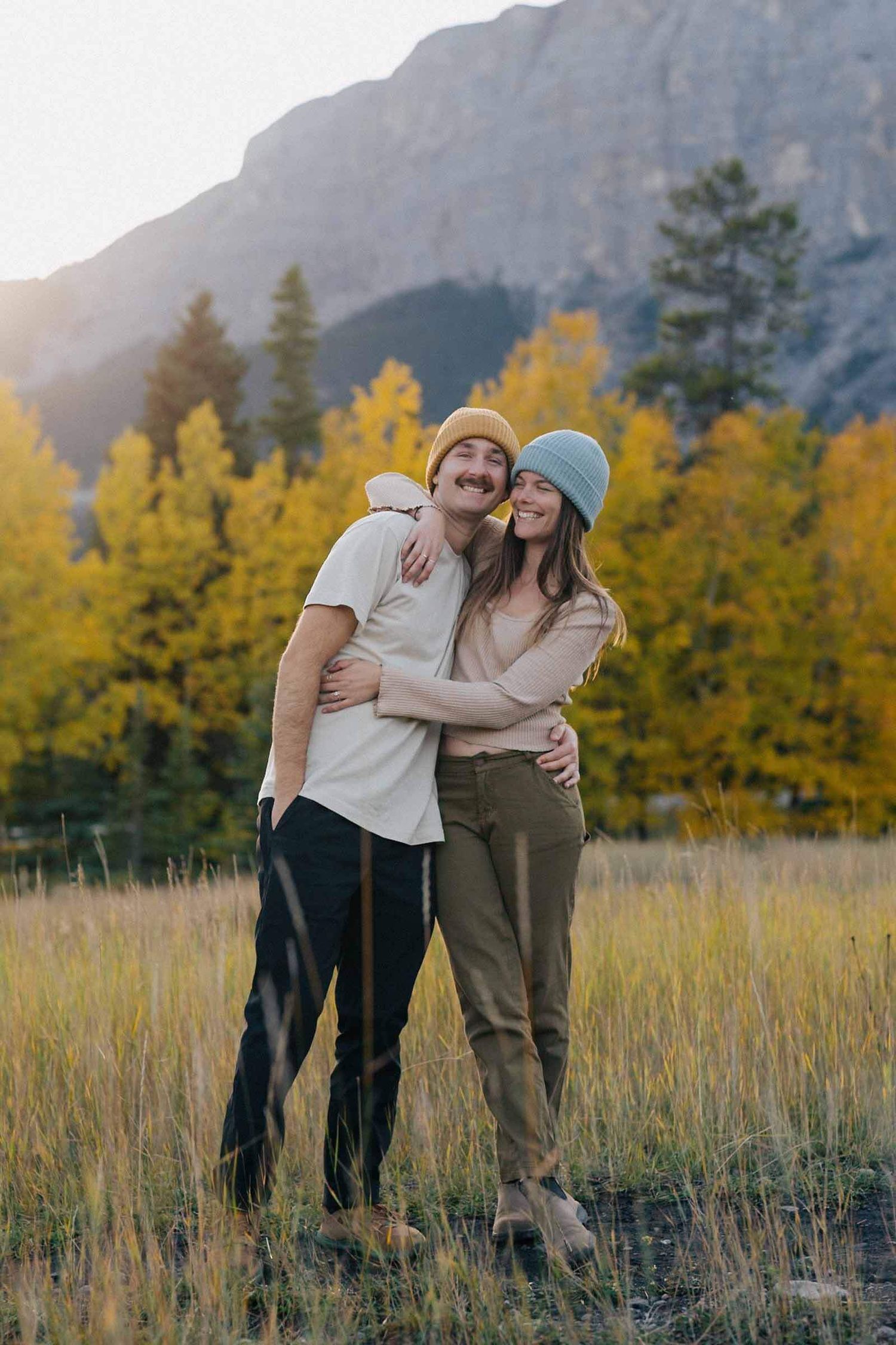 A couple embraces in a meadow with autumn foliage and mountain views at sunset.