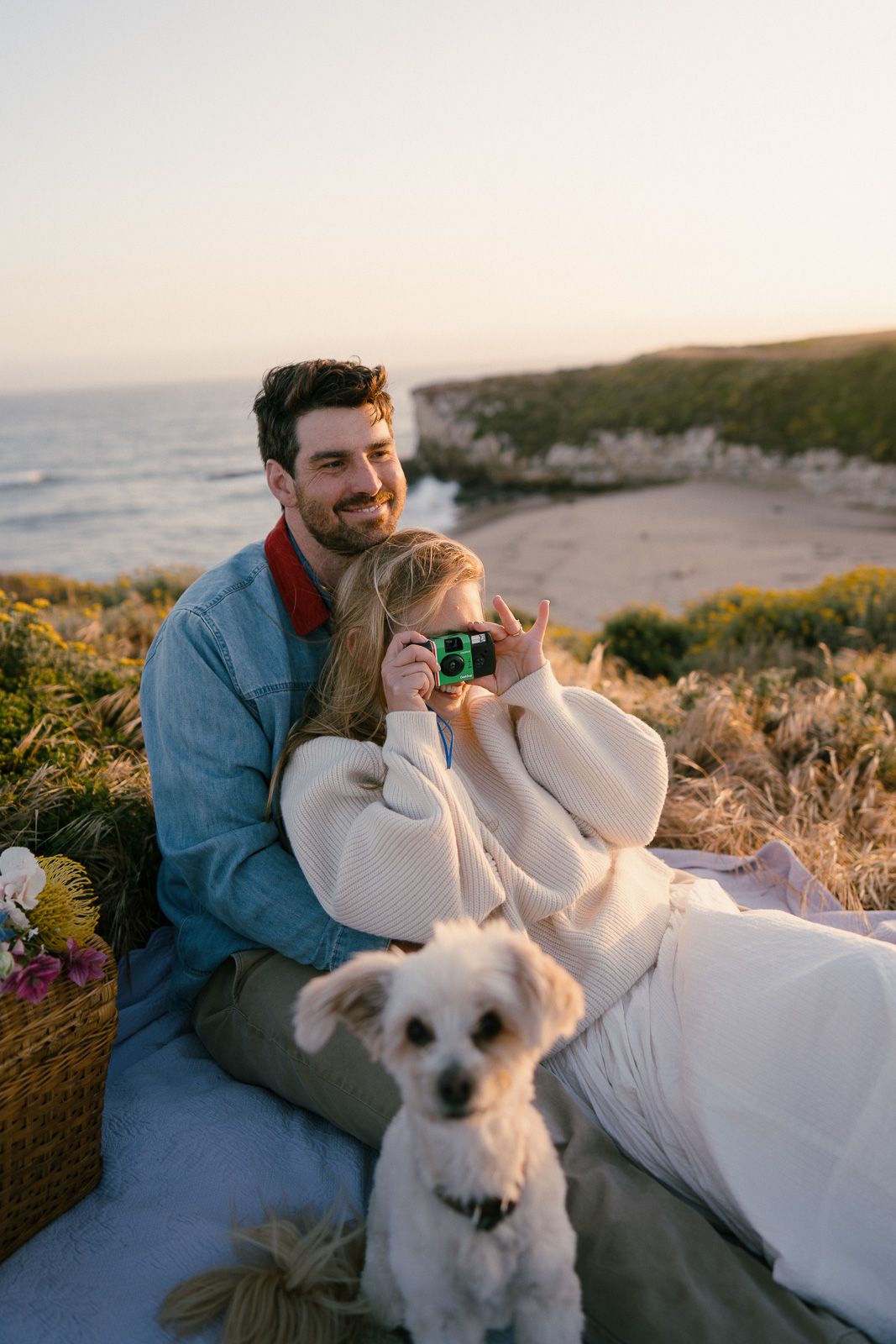 Couple relaxes on coastal cliff during sunset with small white dog and picnic basket at scenic overlook.