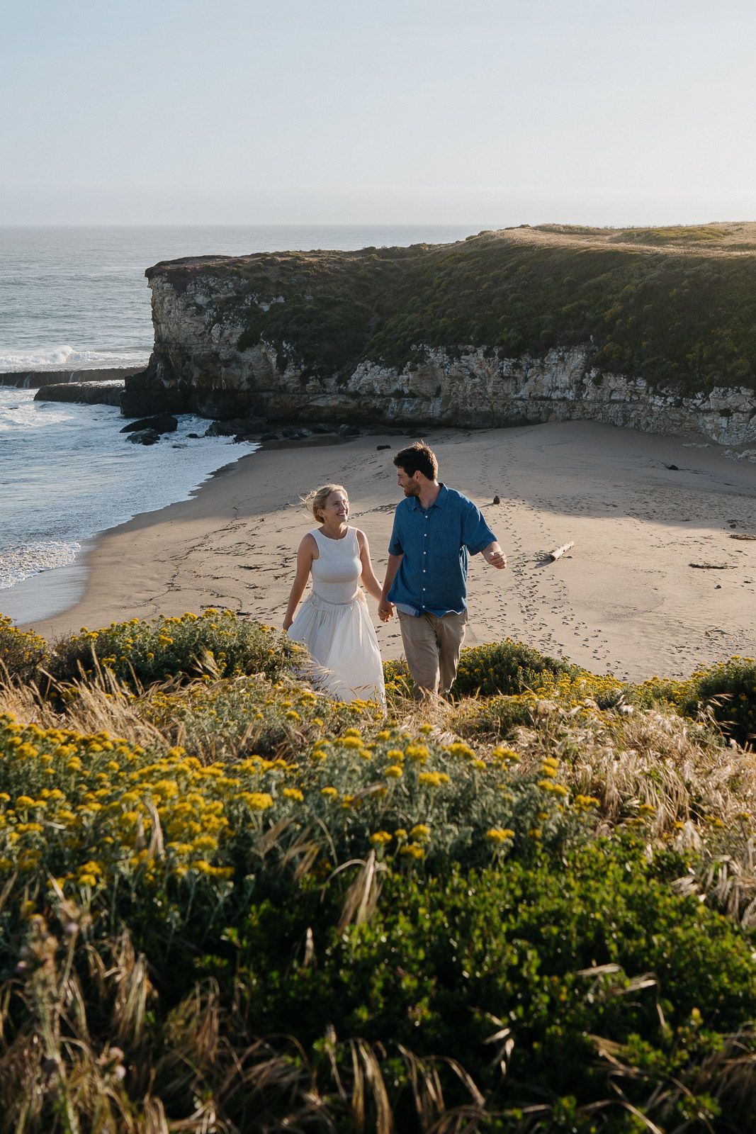 Couple walking along a coastal cliffside path among yellow wildflowers with ocean views at sunset.