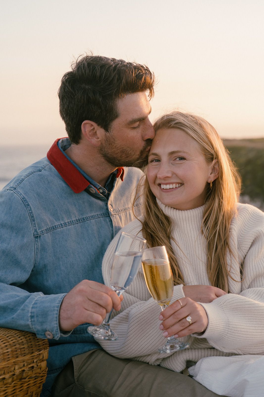 Cozy couple shares champagne while sitting on blanket at sunset during romantic beachside picnic.