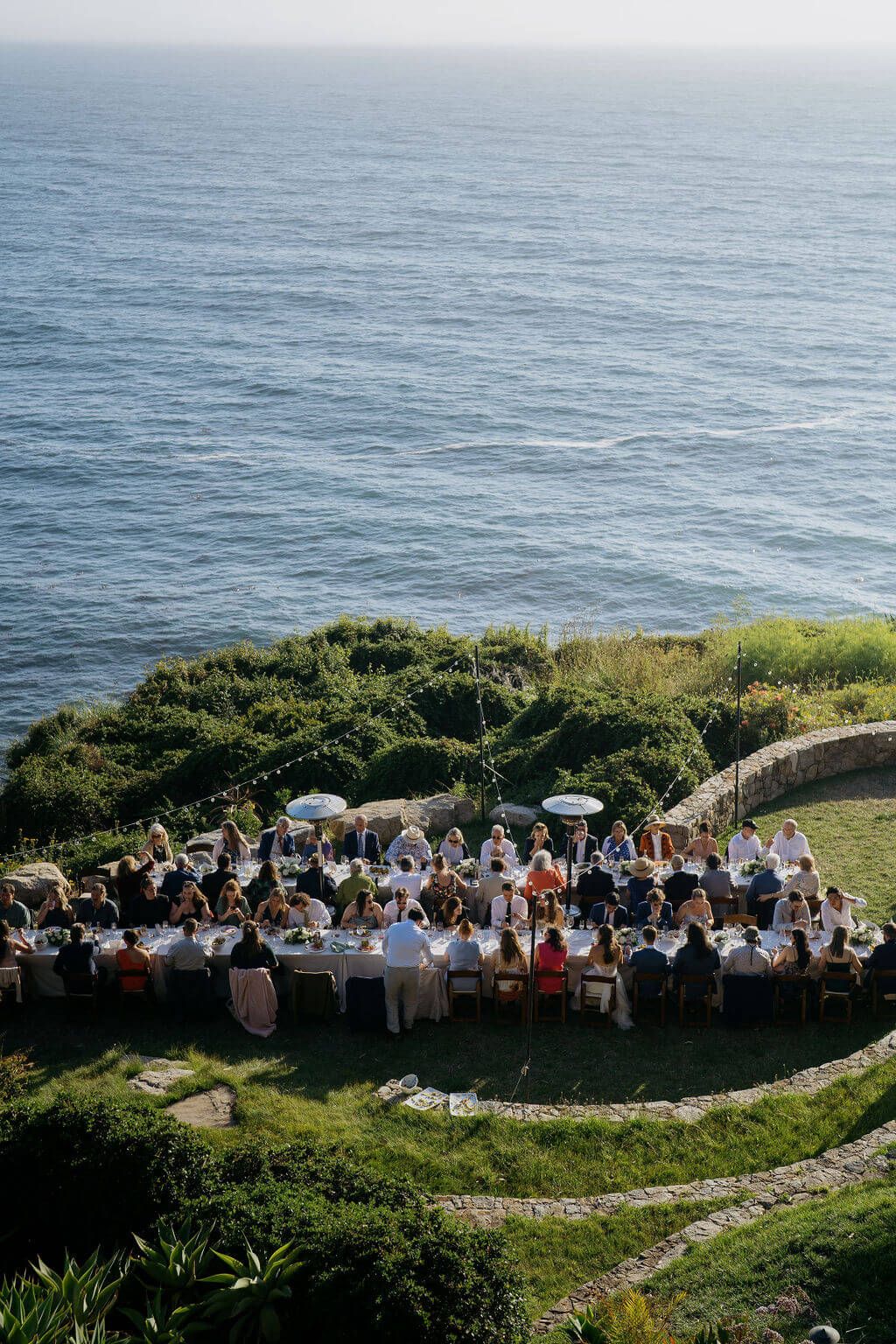 Aerial view of an outdoor wedding reception by the ocean cliff with guests seated at long tables surrounded by grass.