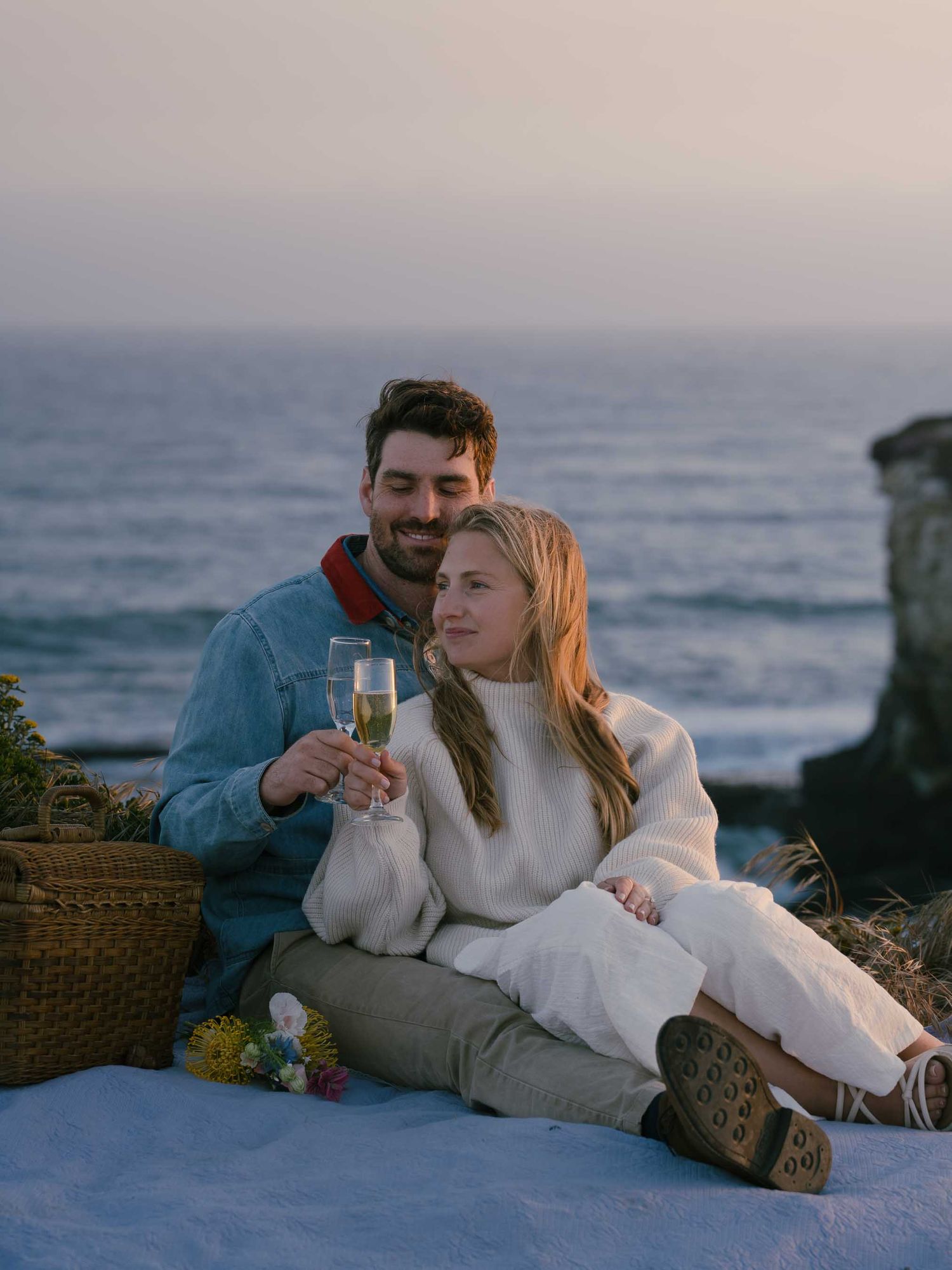 Couple enjoying champagne during romantic sunset picnic by the ocean on a blanket with picnic basket.
