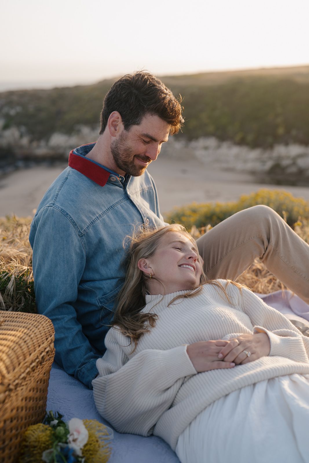 Couple enjoying sunset picnic on hillside with wicker basket and blanket in warm golden light.