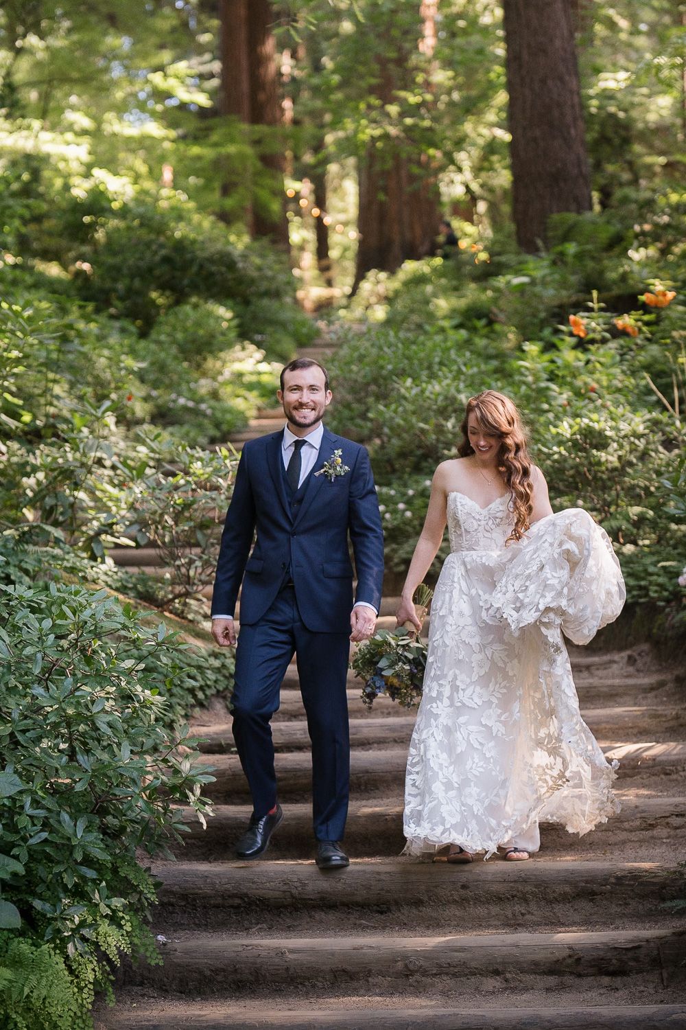 A bride in a lace dress and groom in navy suit walking up forest stairs surrounded by lush greenery.