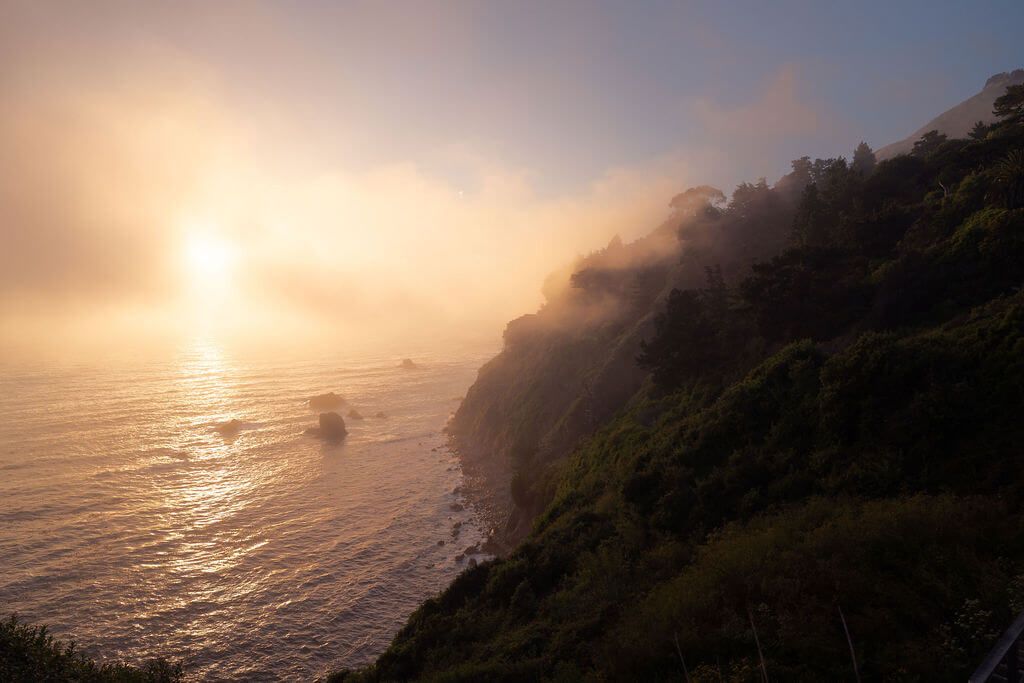 Golden sunset illuminates foggy coastal cliffs along the Pacific Ocean as mist rolls in over the rocky shoreline.